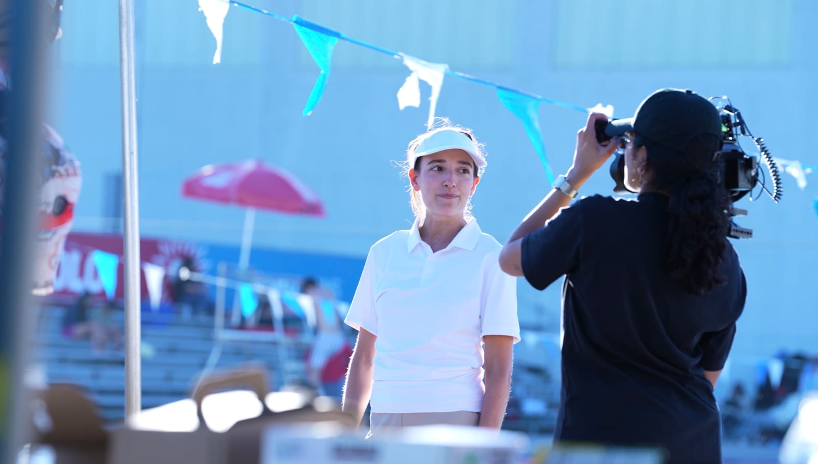 Person in white shirt and cap being photographed by another person with camera, outdoor setting with umbrellas and decorations