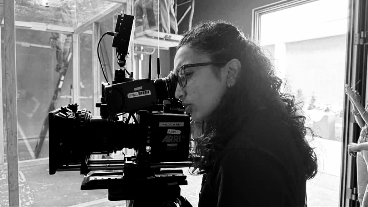Person with curly hair and glasses looking through a microscope, in a room with large windows and equipment.