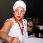 Older woman wearing a white headwrap and dress, holding a wooden mallet, with a large drum in front of her.