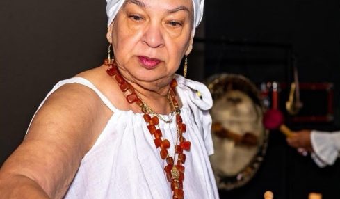 Older woman wearing a white headwrap and dress, holding a wooden mallet, with a large drum in front of her.