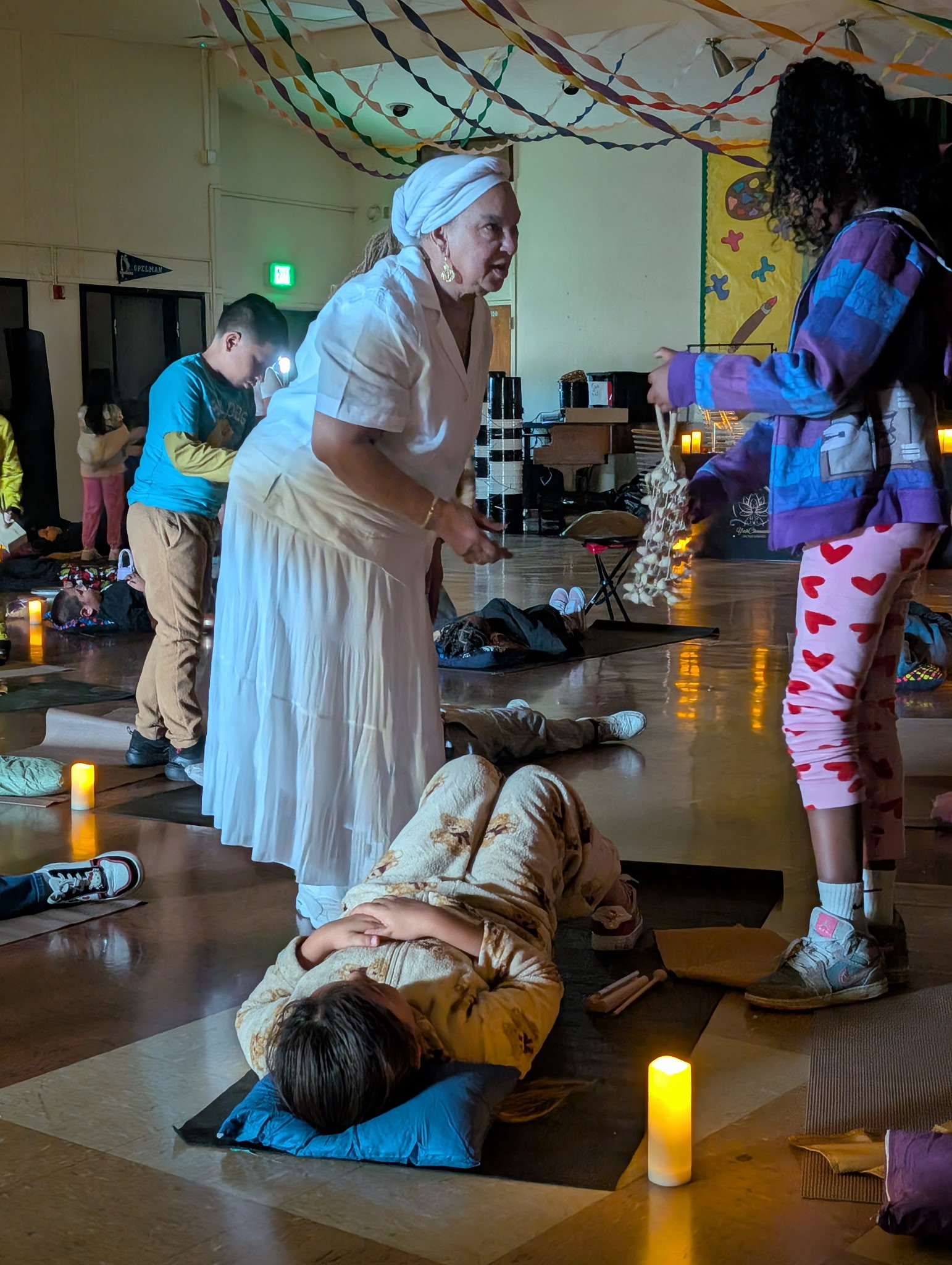 A woman in a white dress and headscarf kneels beside a person lying on the floor with a pillow, in a decorated room.