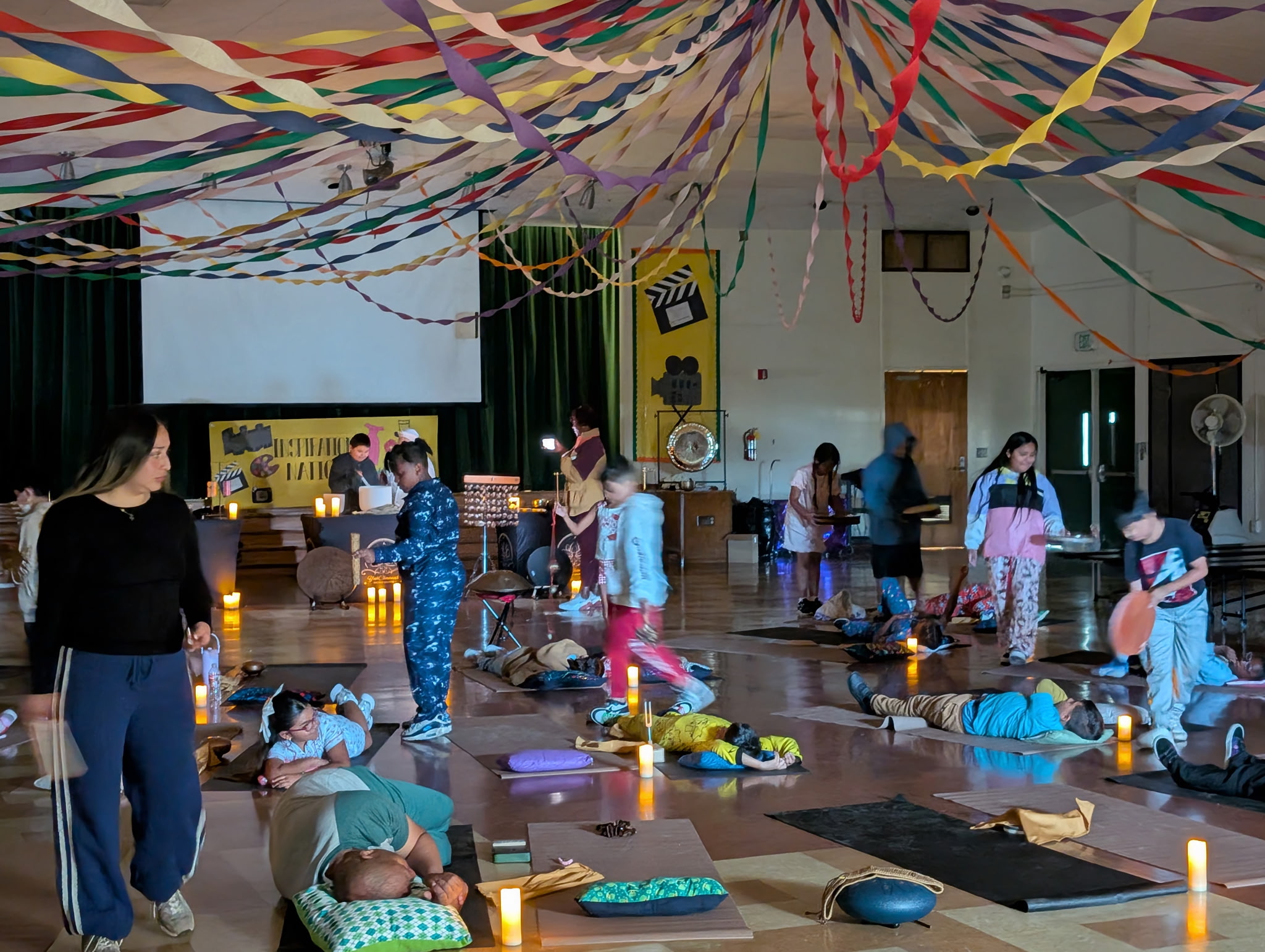 Children and adults participate in a yoga class in a decorated hall with streamers and candles, some lying on mats.