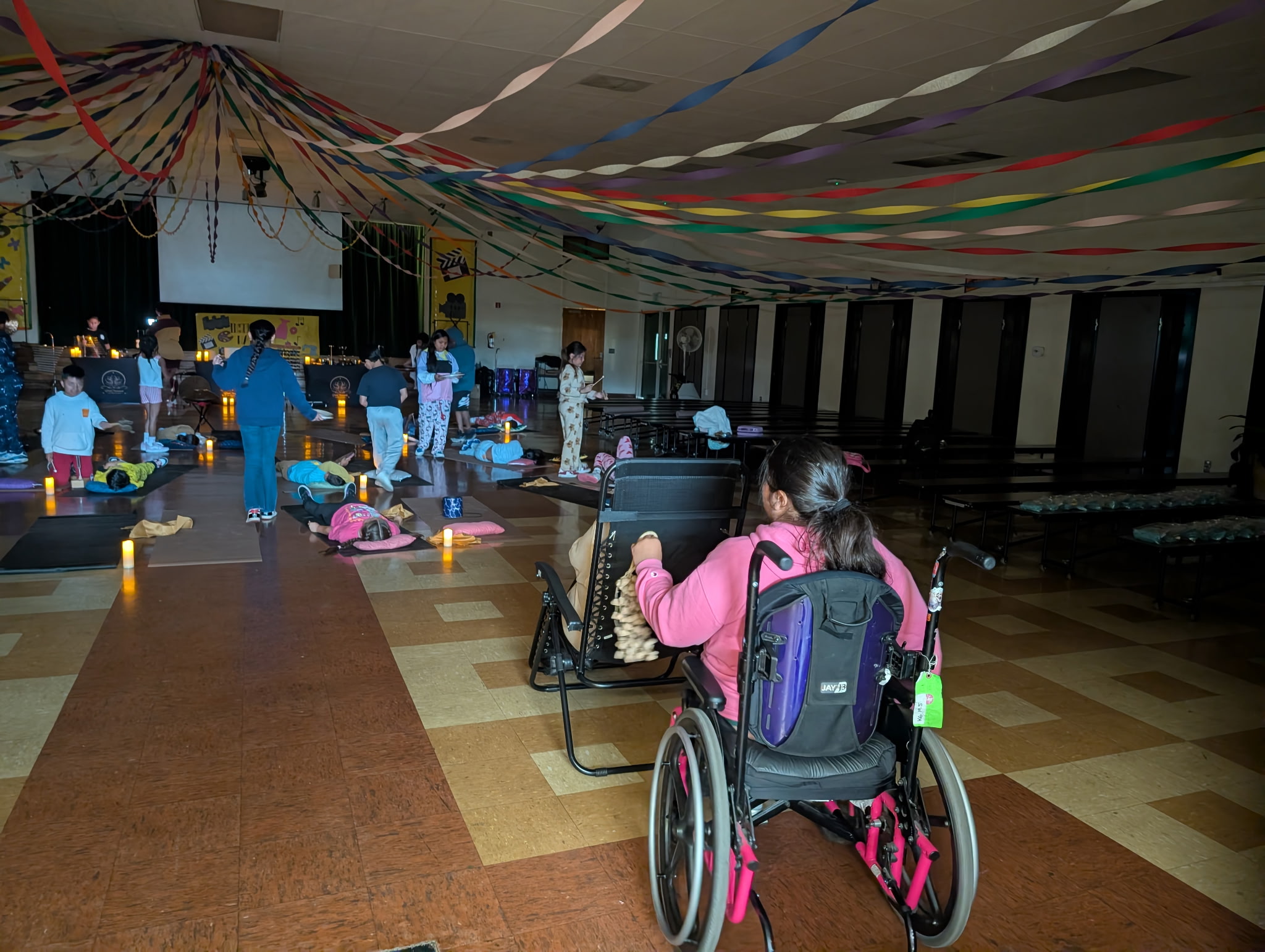 Person in wheelchair facing a stage with people and decorations in a large indoor event space.