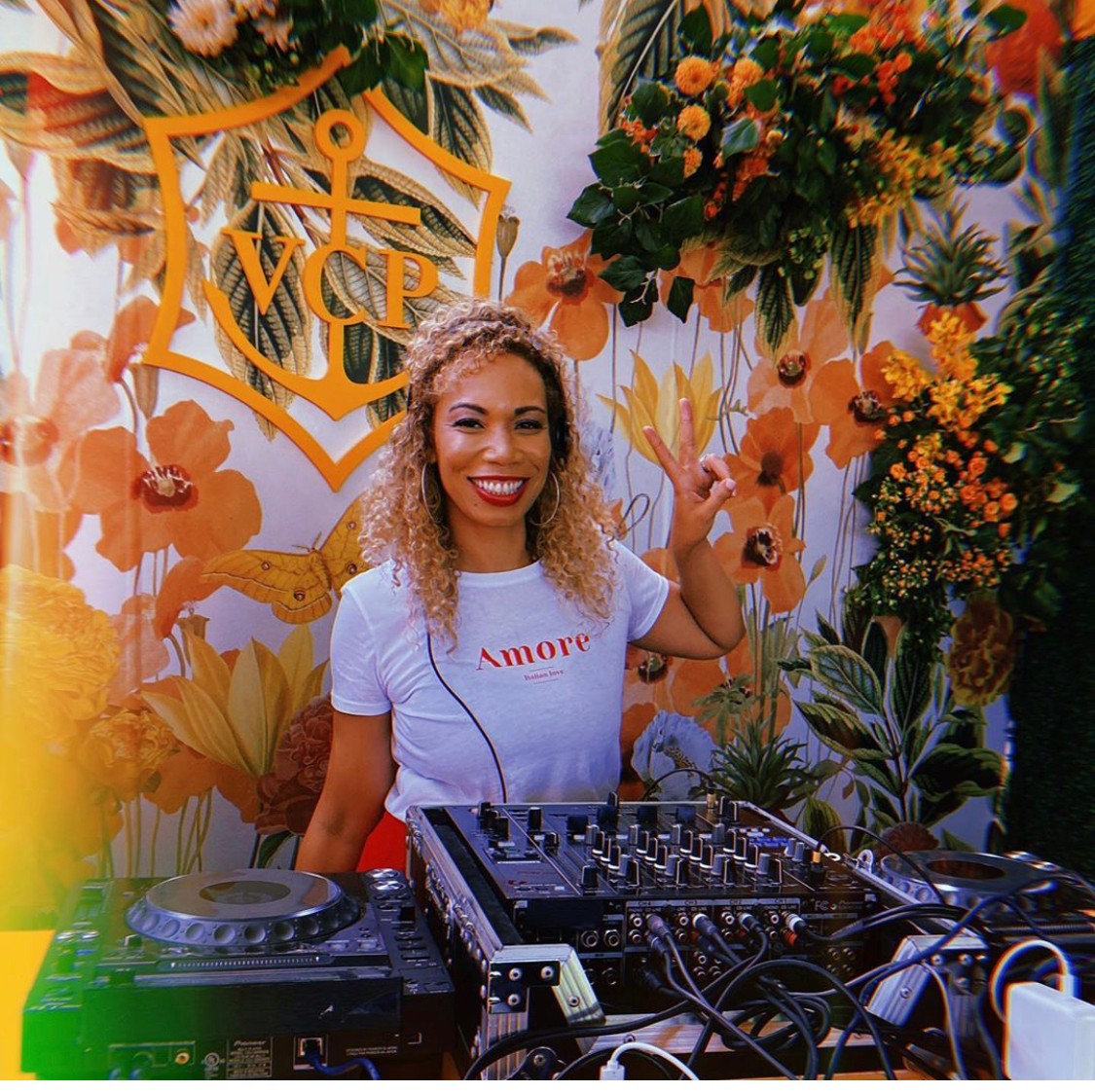 Smiling woman with curly hair making a peace sign at a DJ booth surrounded by flowers and a VIP sign.
