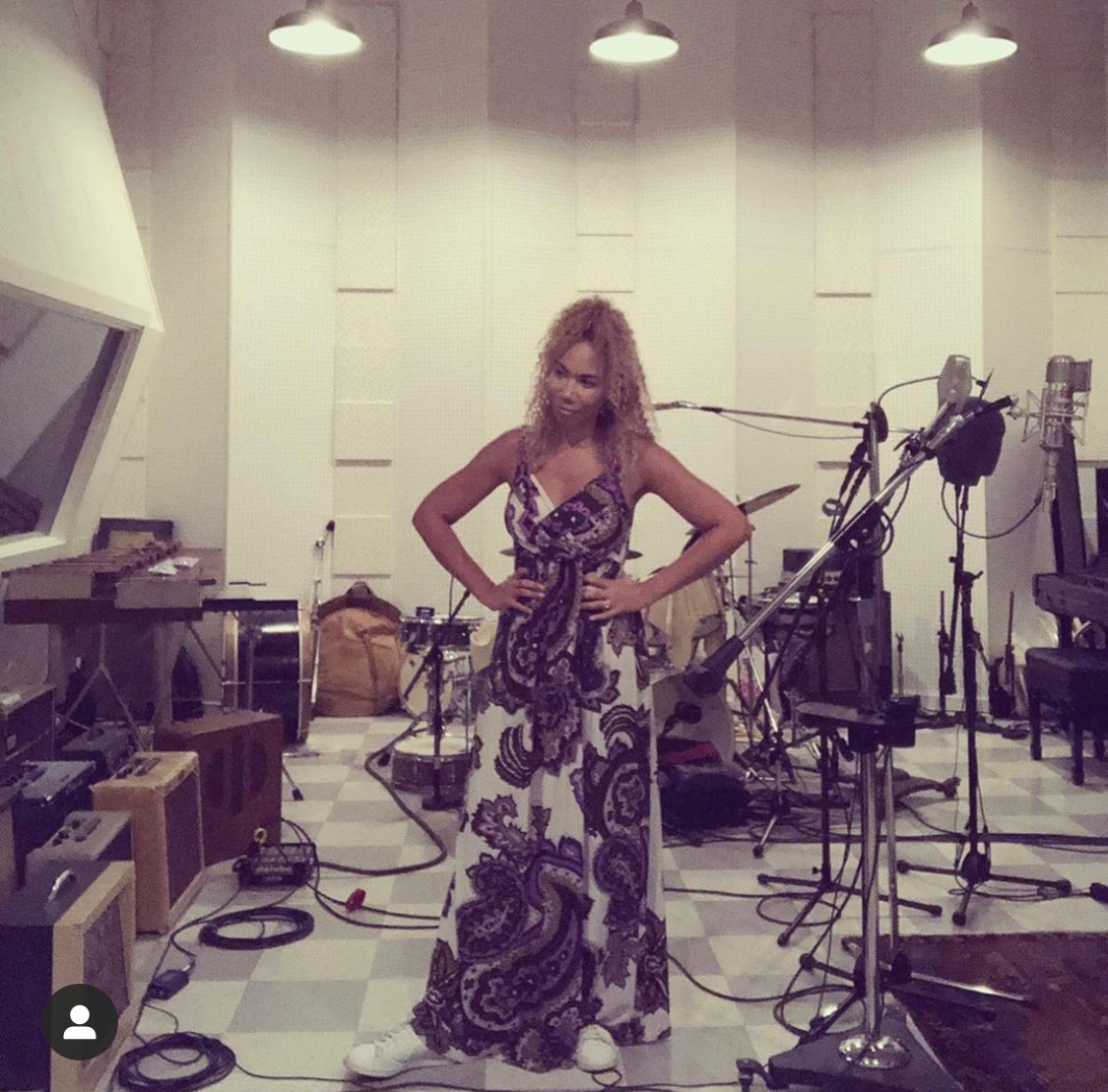 Woman in a patterned dress stands in a music studio with microphones and equipment, hands on hips, looking down.