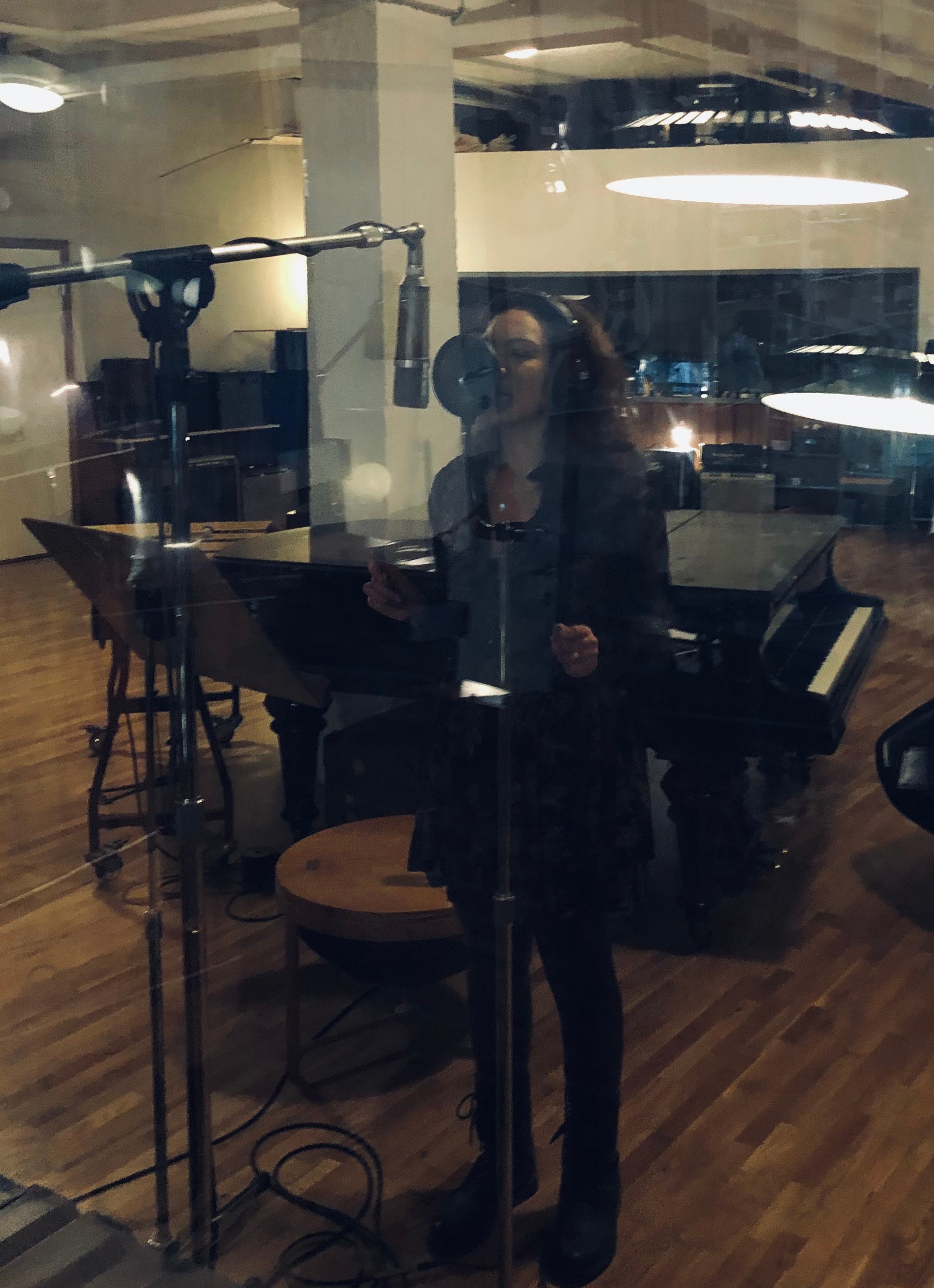 Person standing in recording studio with microphone, music stand, and piano, reflected in glass window, dim lighting, wooden floor.