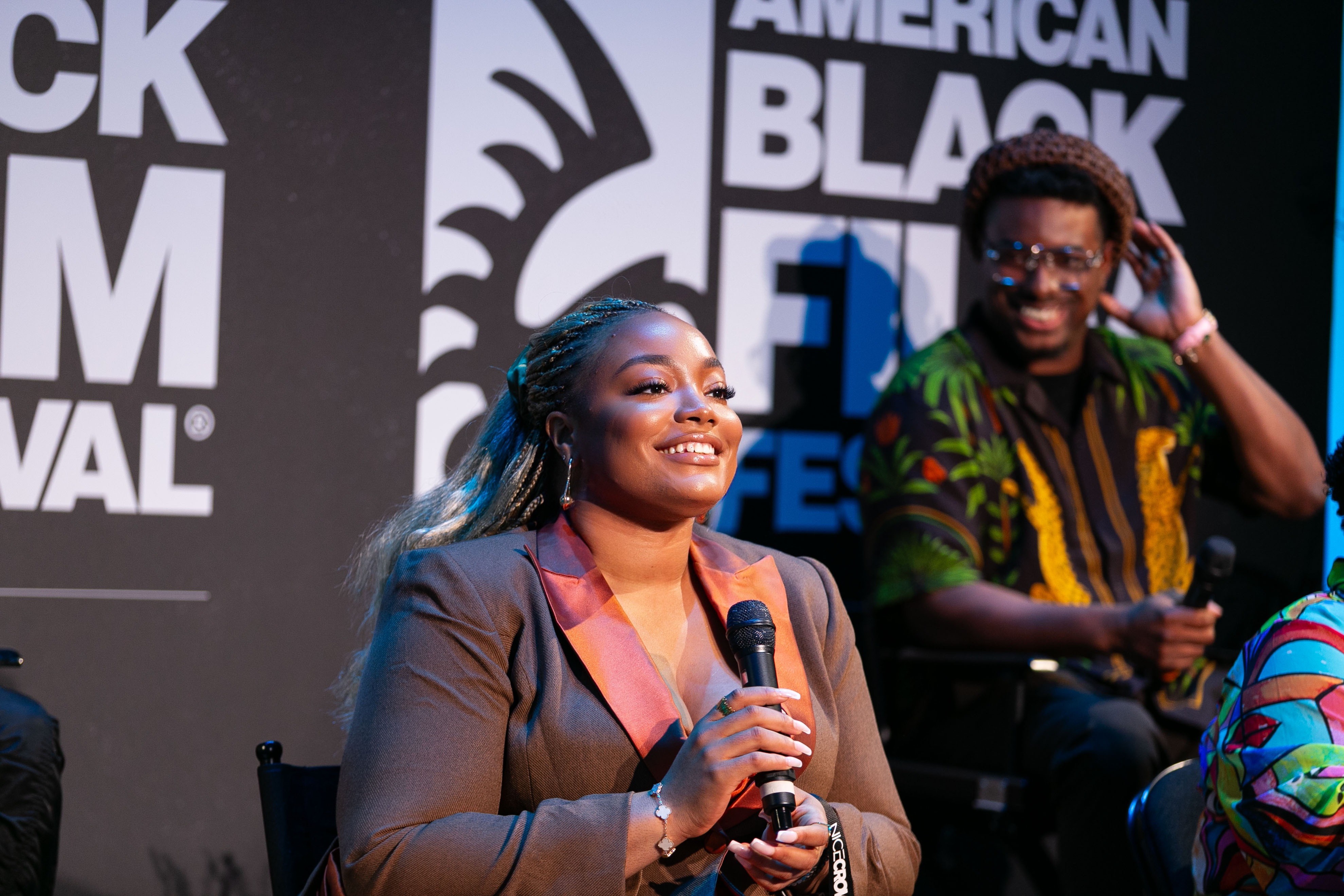 Two people on stage, one woman speaking into a microphone, smiling, and a man smiling in the background, with a black and white backdrop.