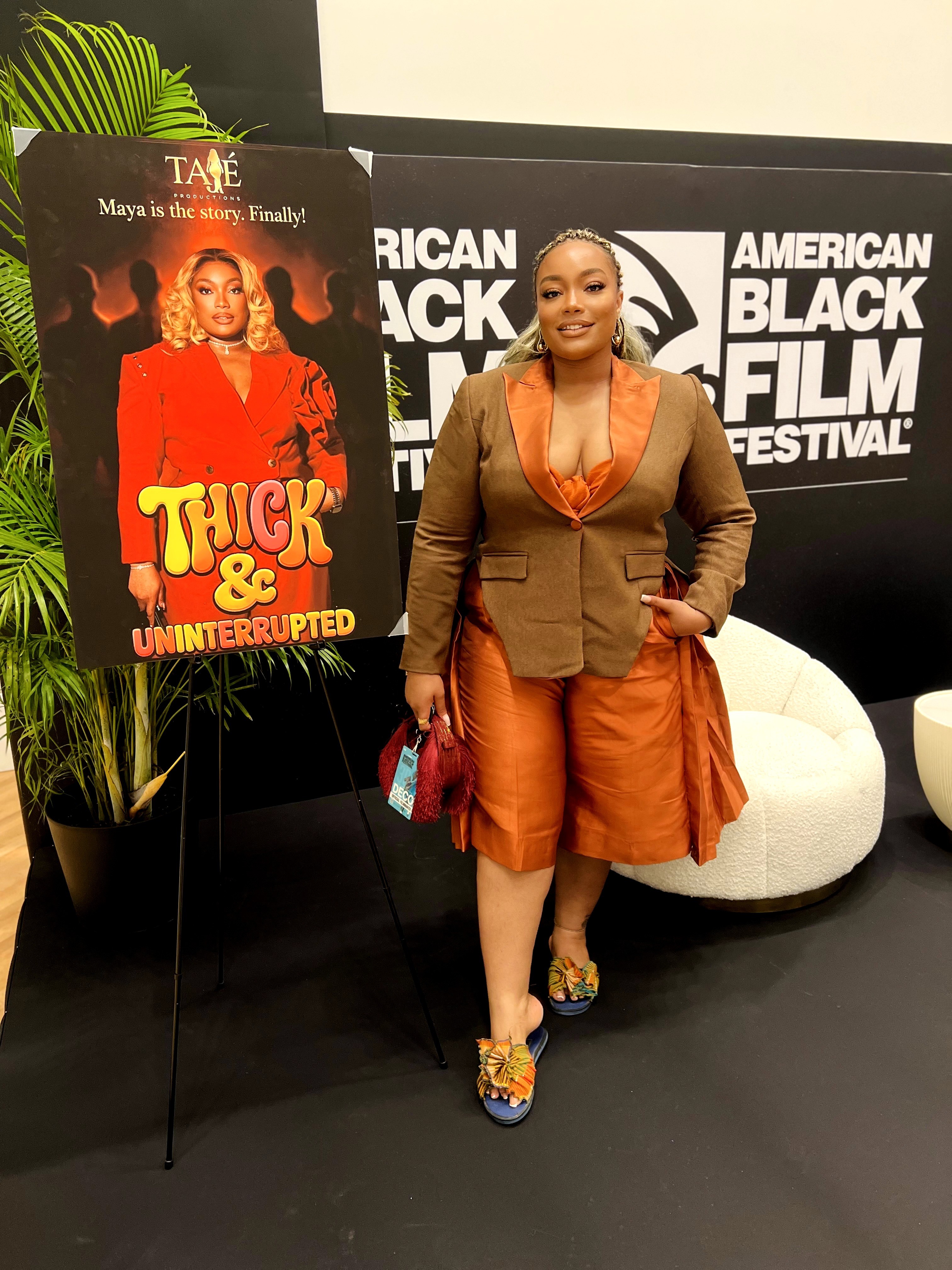 Woman in orange dress and brown blazer standing in front of a sign at a film festival, with a poster on an easel nearby.