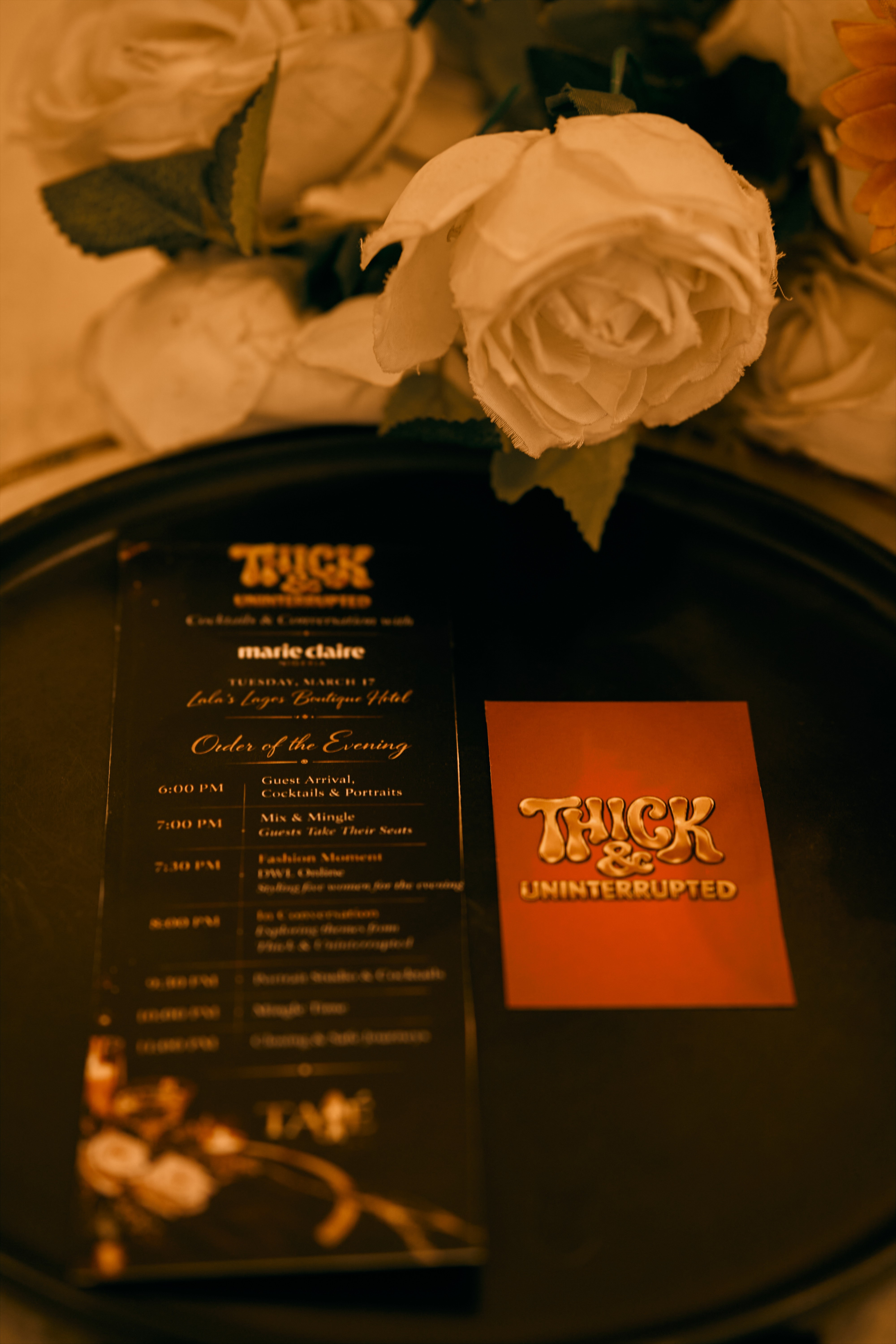 White rose with green leaves on a black tray, with a red and black card and a black menu nearby.