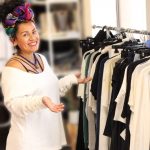 Smiling woman with colorful headscarf shopping for clothes in a store, pointing at a rack of various garments.