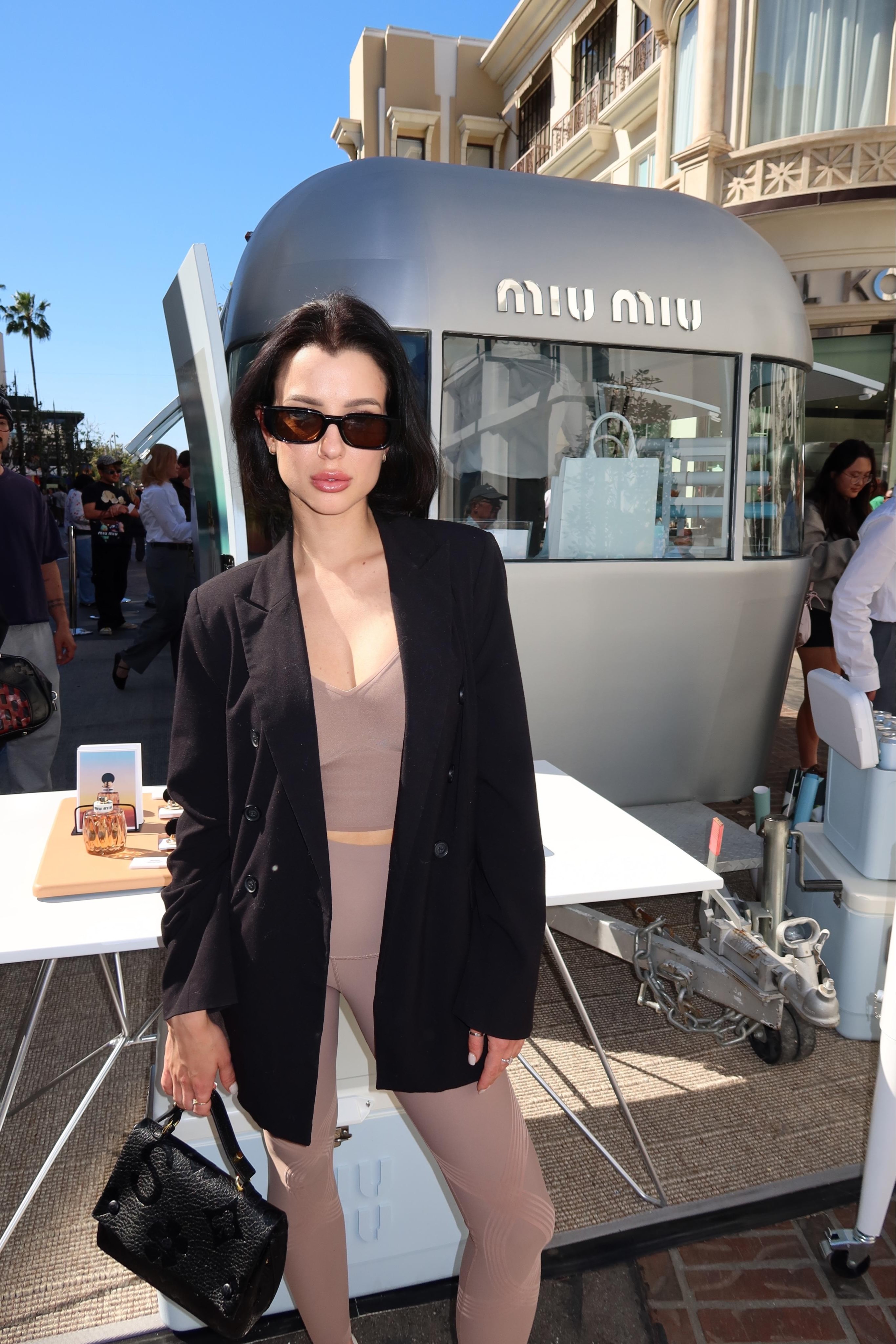 Woman with dark hair and sunglasses stands outdoors in front of a modern tram, wearing a black blazer and beige outfit.