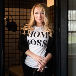 Woman with long blonde hair wearing a white T-shirt with 'MOM BOSS' text, standing in doorway holding headphones, smiling.