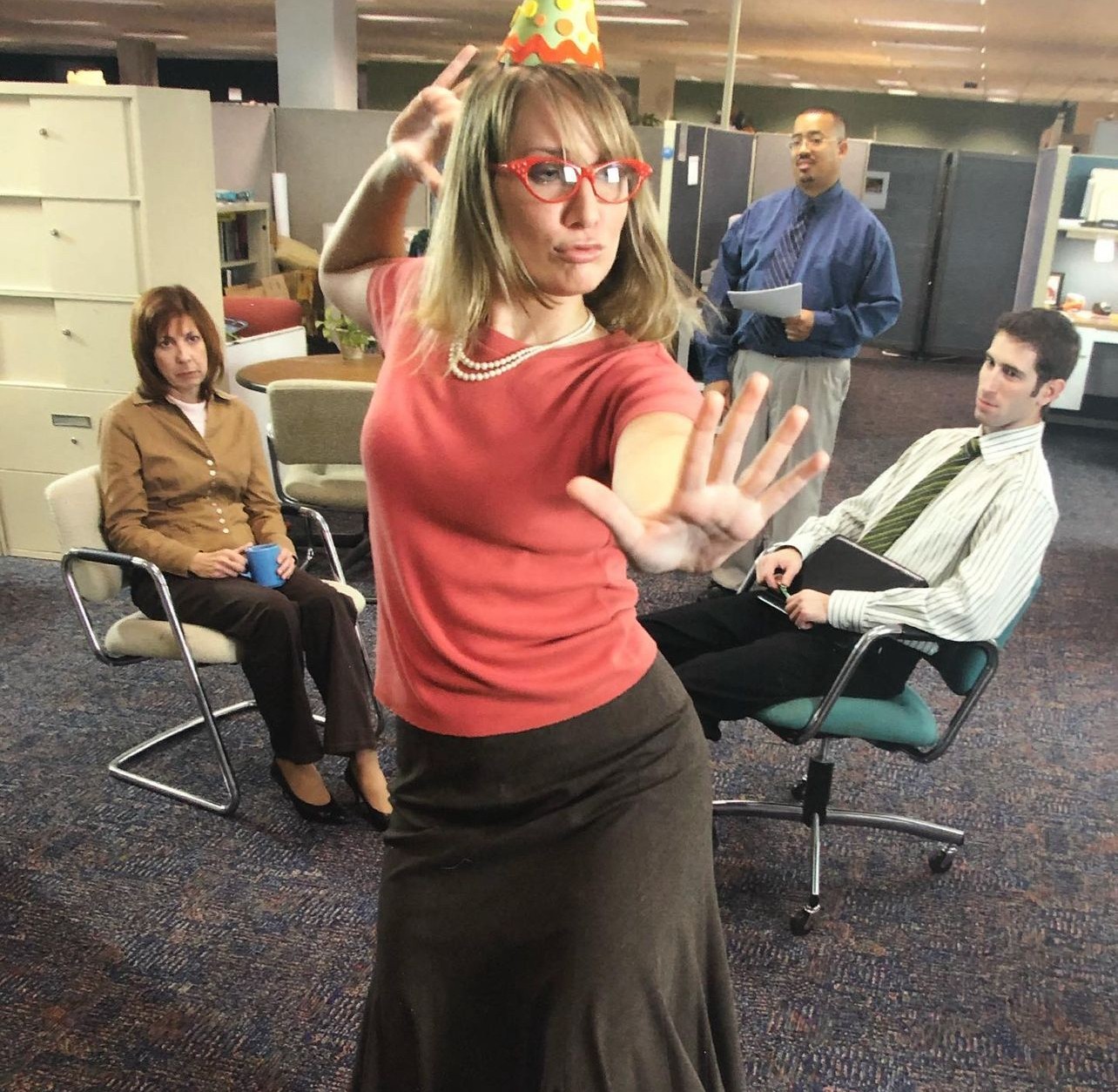 Woman in party hat and red glasses raises hand in office setting, three people seated behind her.