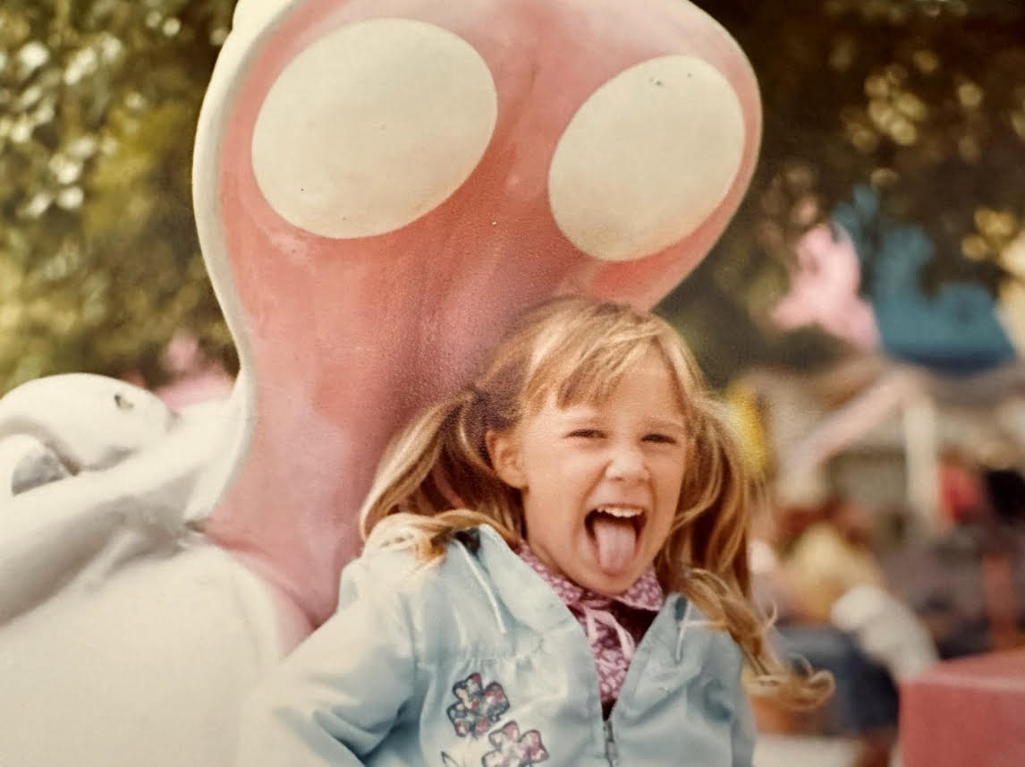 Young girl laughing with mouth open, standing near a large pink and white sculpture outdoors, blurred background.