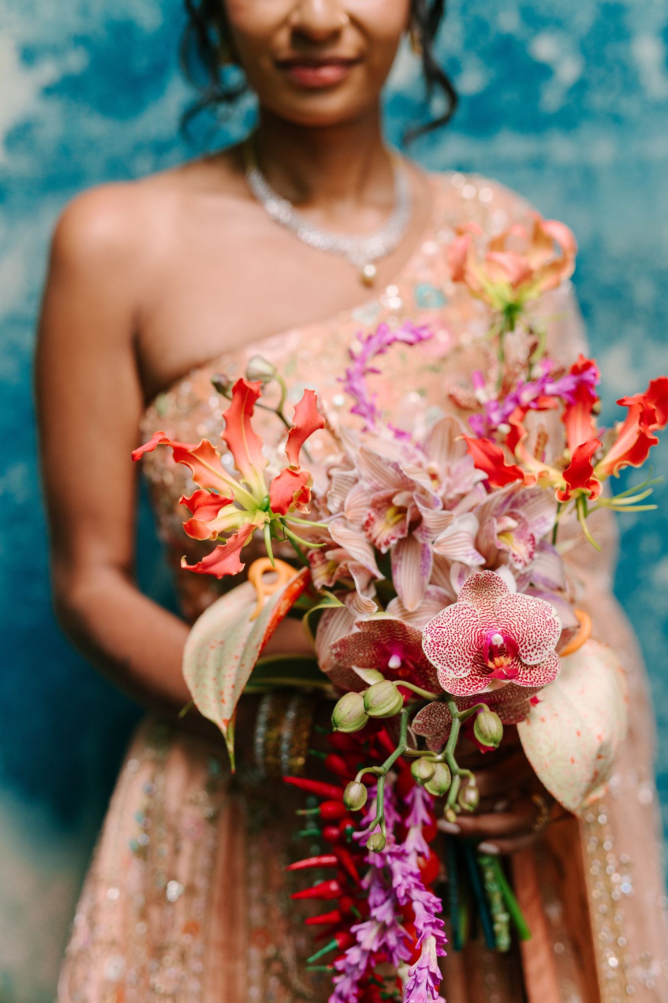 Person holding a colorful bouquet of flowers, wearing a strapless dress and necklace, with a blue background.