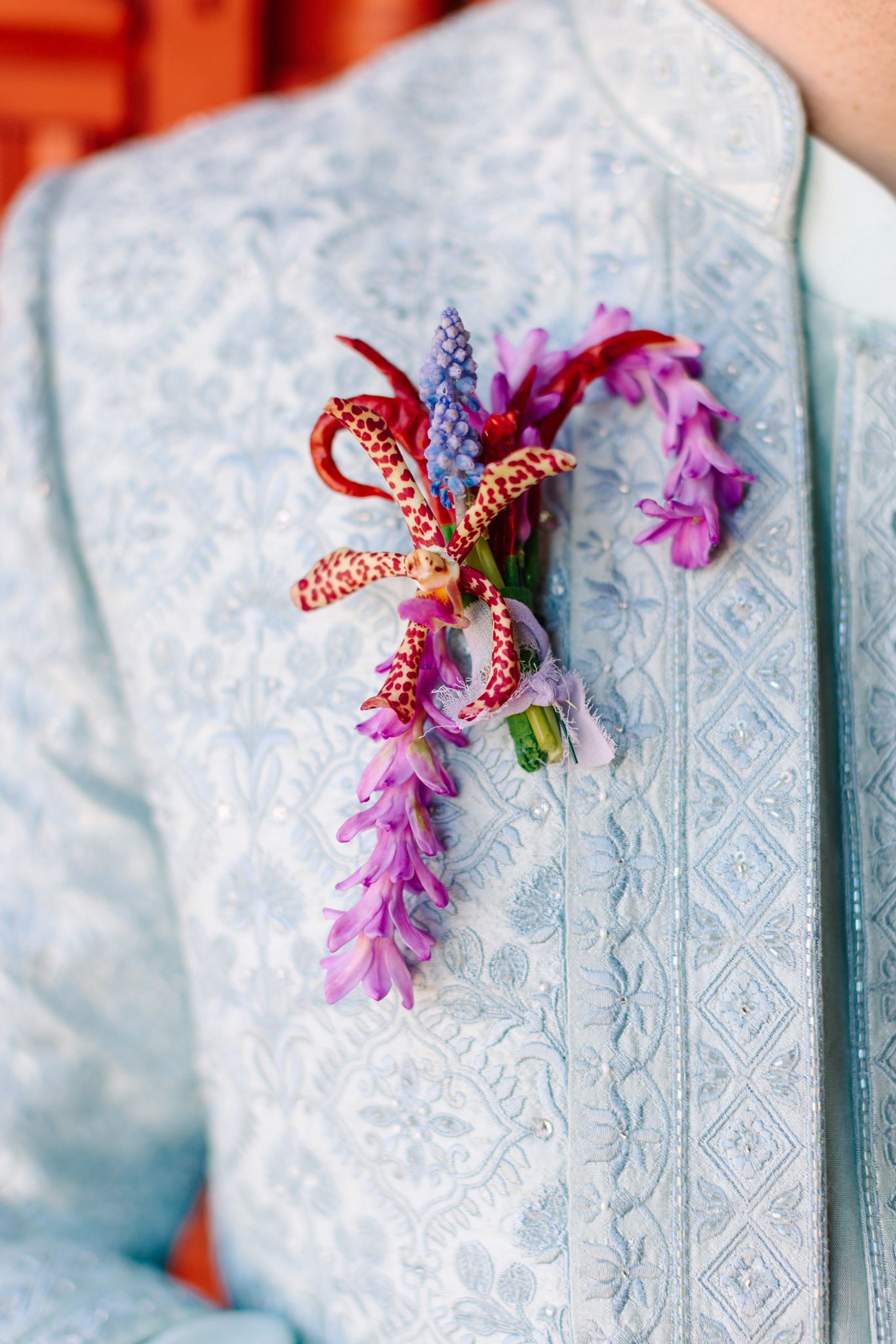 Close-up of a floral boutonniere on a light blue patterned suit jacket, featuring purple and pink flowers with decorative ribbons.