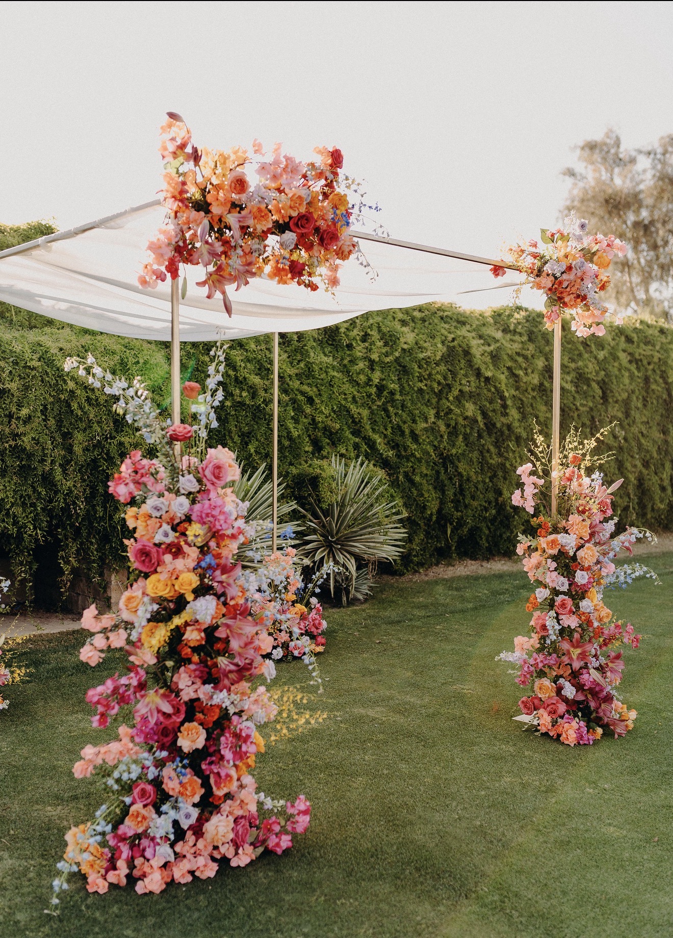 Flower arch with pink, orange, and yellow flowers on a grassy outdoor setting, decorated for an event.
