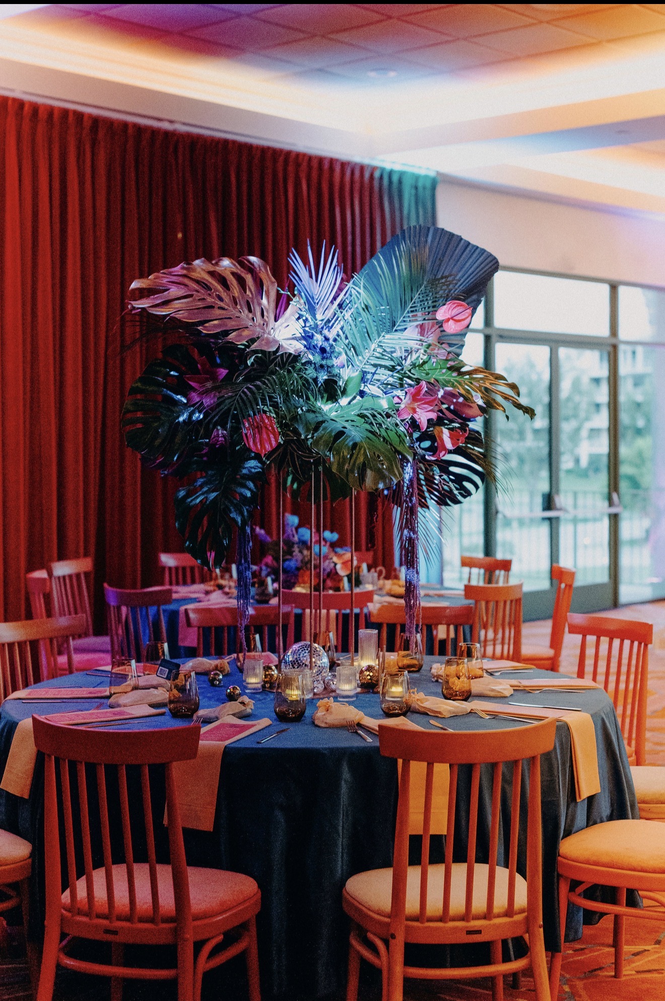 Elegant dining table with a large floral centerpiece, set with plates, glasses, and cutlery, in a room with red curtains and large windows.