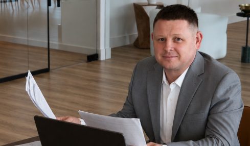 Man in gray suit sitting at desk with laptop and papers in modern office, glass walls, wooden floor, and plants.