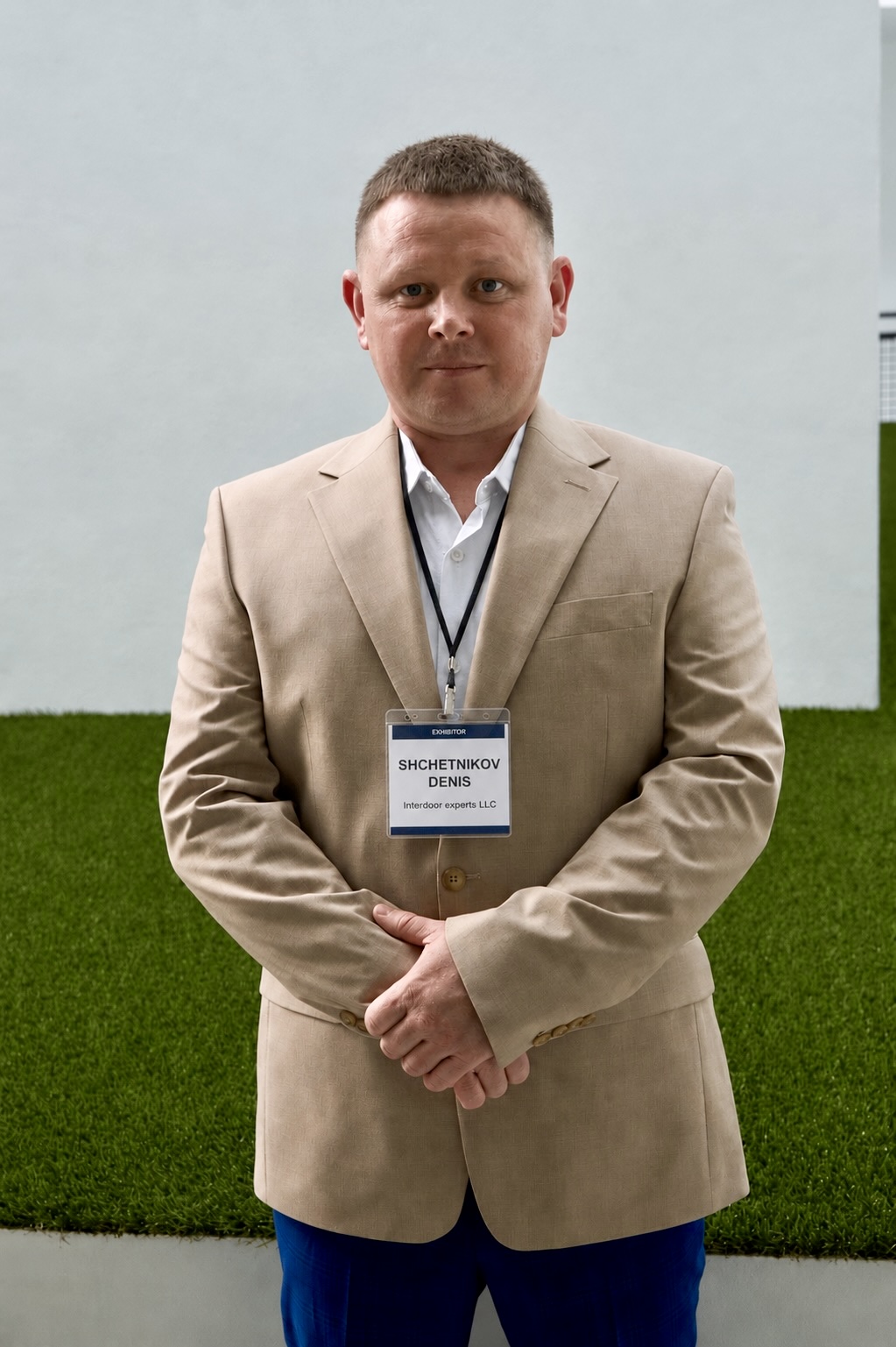Man in beige suit with name badge standing outdoors against a background of grass and a wall.