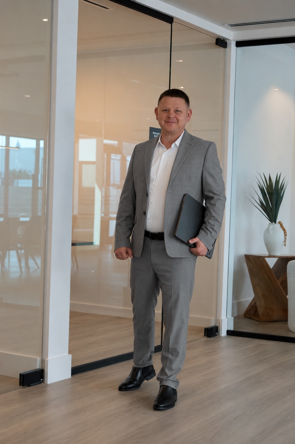 Man in gray suit holding a folder, standing in a modern office space with glass walls and wooden flooring.