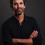 Man with dark hair and beard, wearing a black shirt, crossing arms, looking at camera, neutral expression.