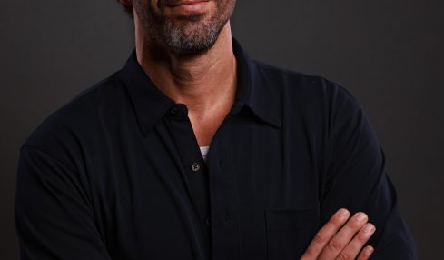 Man with dark hair and beard, wearing a black shirt, crossing arms, looking at camera, neutral expression.