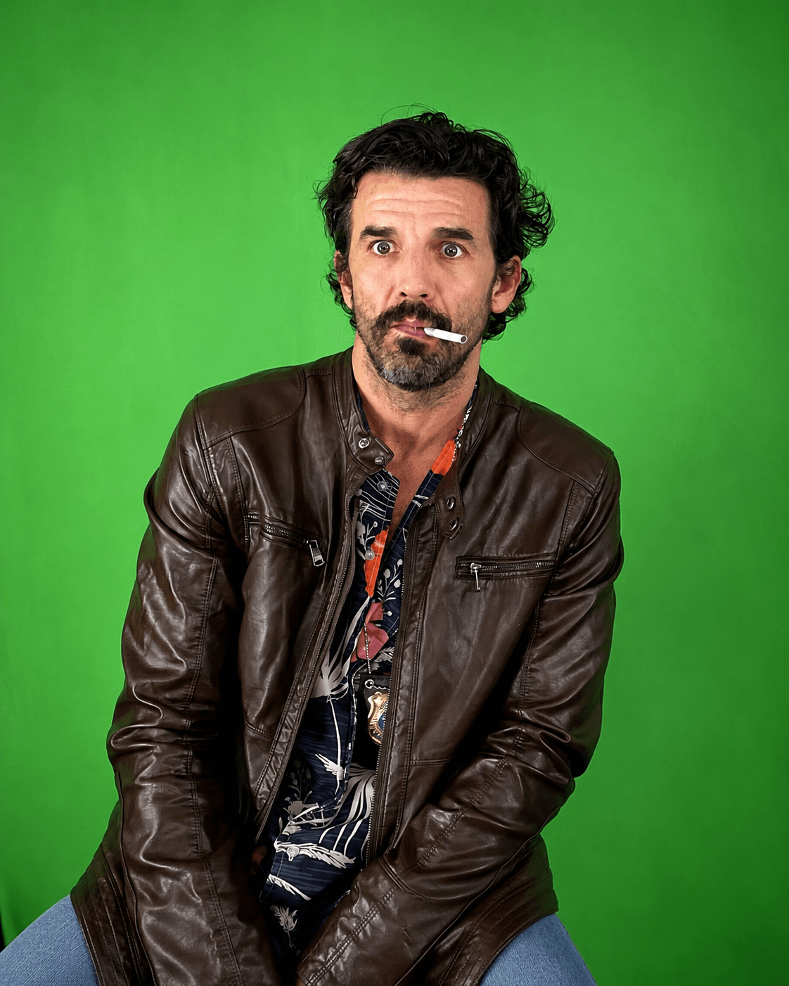 Man with dark curly hair and beard, wearing a leather jacket, standing against a green background.