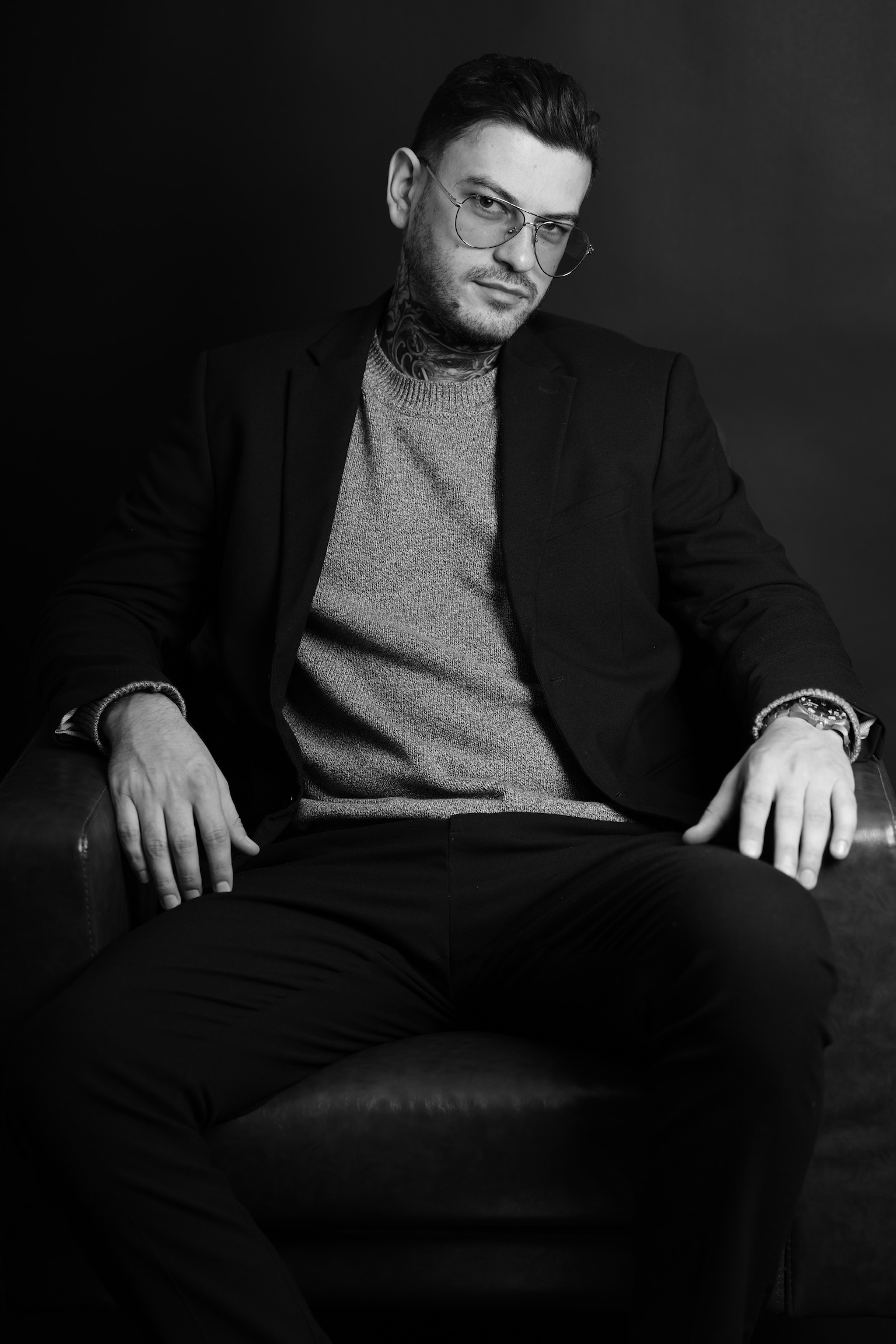 Man with glasses and styled hair sitting in a chair, wearing a blazer and sweater, against a dark background.