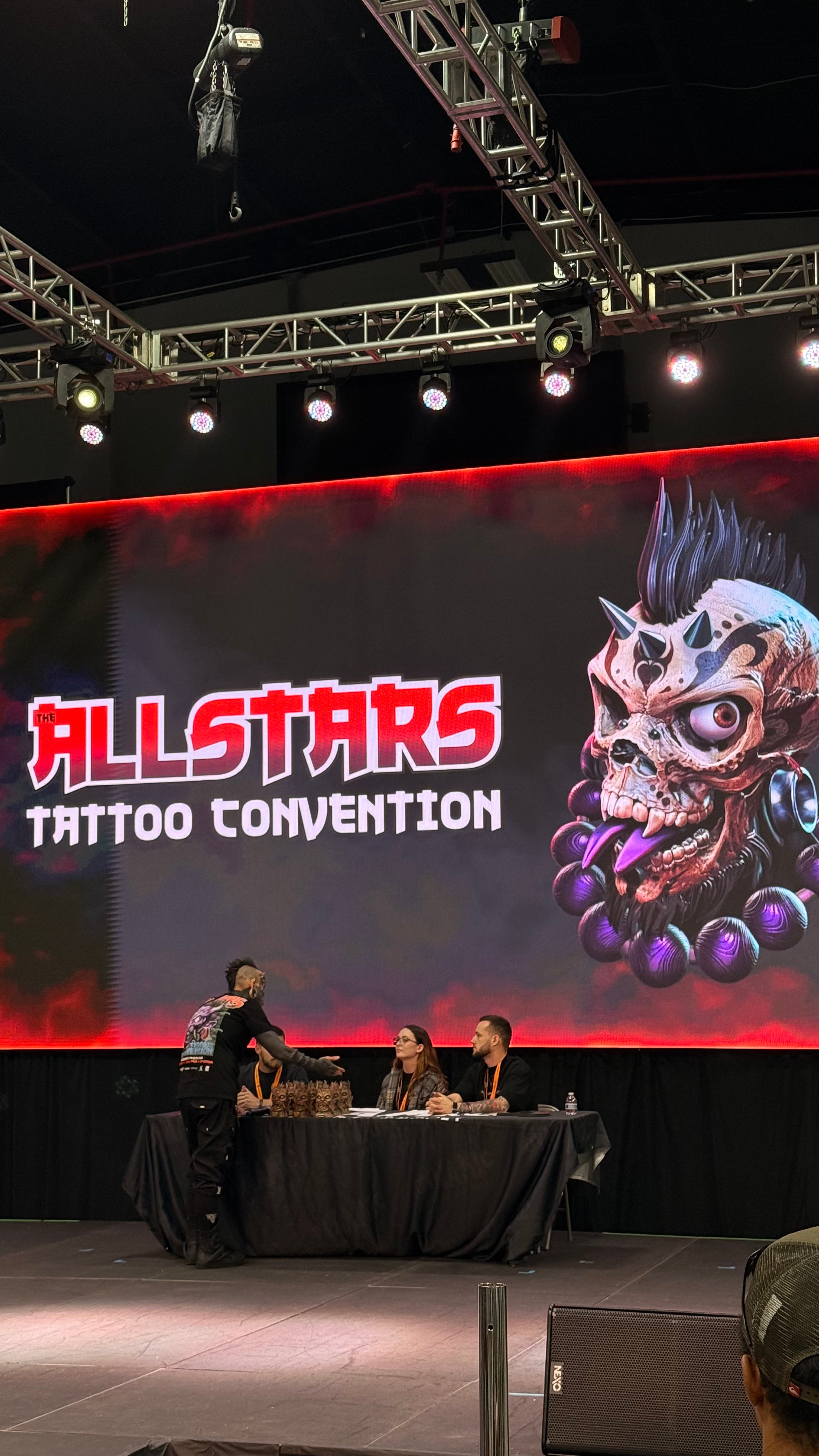 Large screen displays a colorful skull with tattoos and text reading 'ALLSTARS TATTOO CONVENTION'. Three people sit at a table on stage.