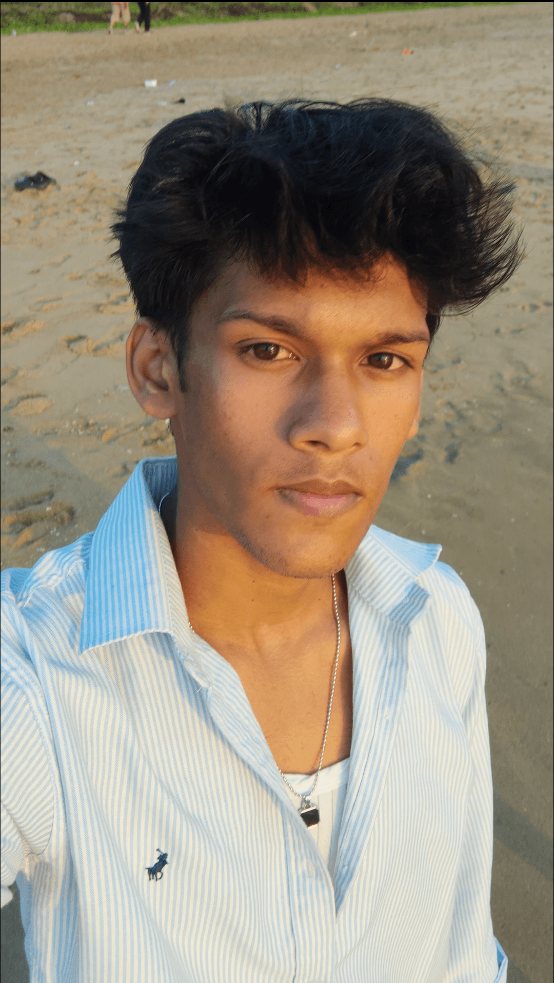 Young man with dark hair and a serious expression, wearing a light blue striped shirt, standing on a sandy area.