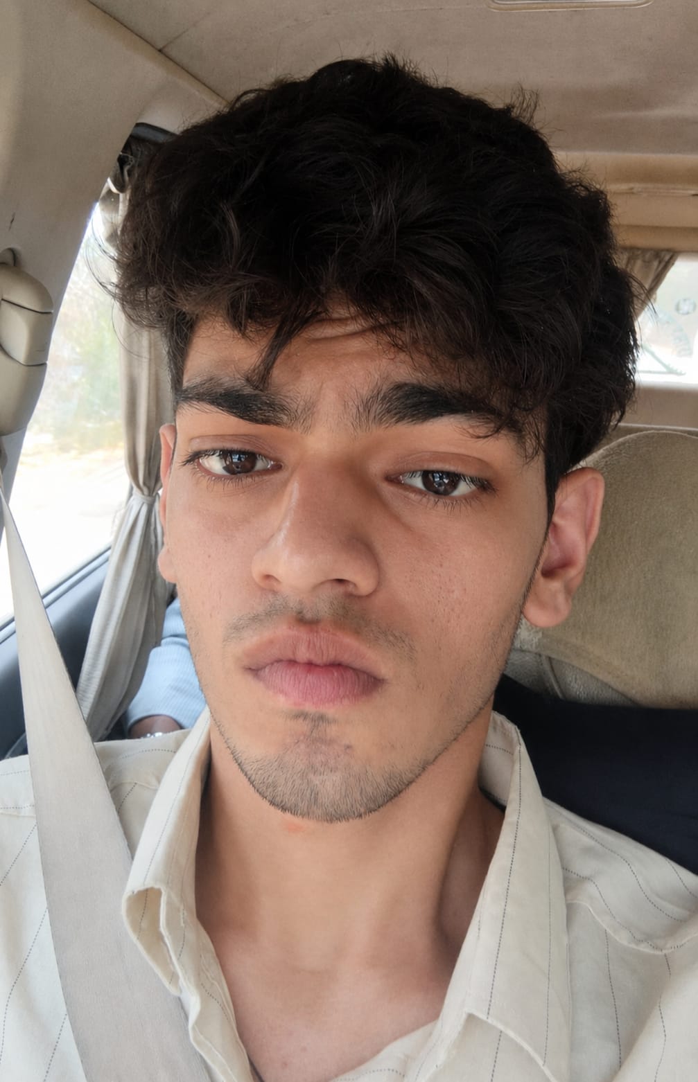 Young man with dark, curly hair and light skin, wearing a light-colored shirt, sitting in a car.