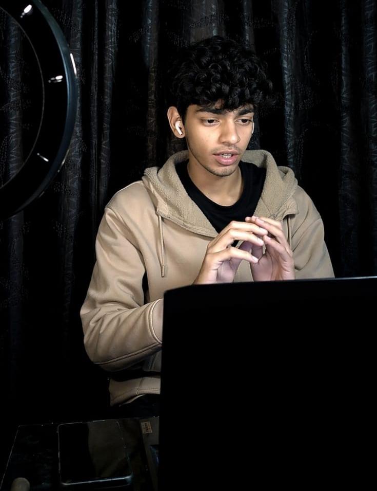 Young person with curly hair using a laptop, dark background, black curtain, focused expression.