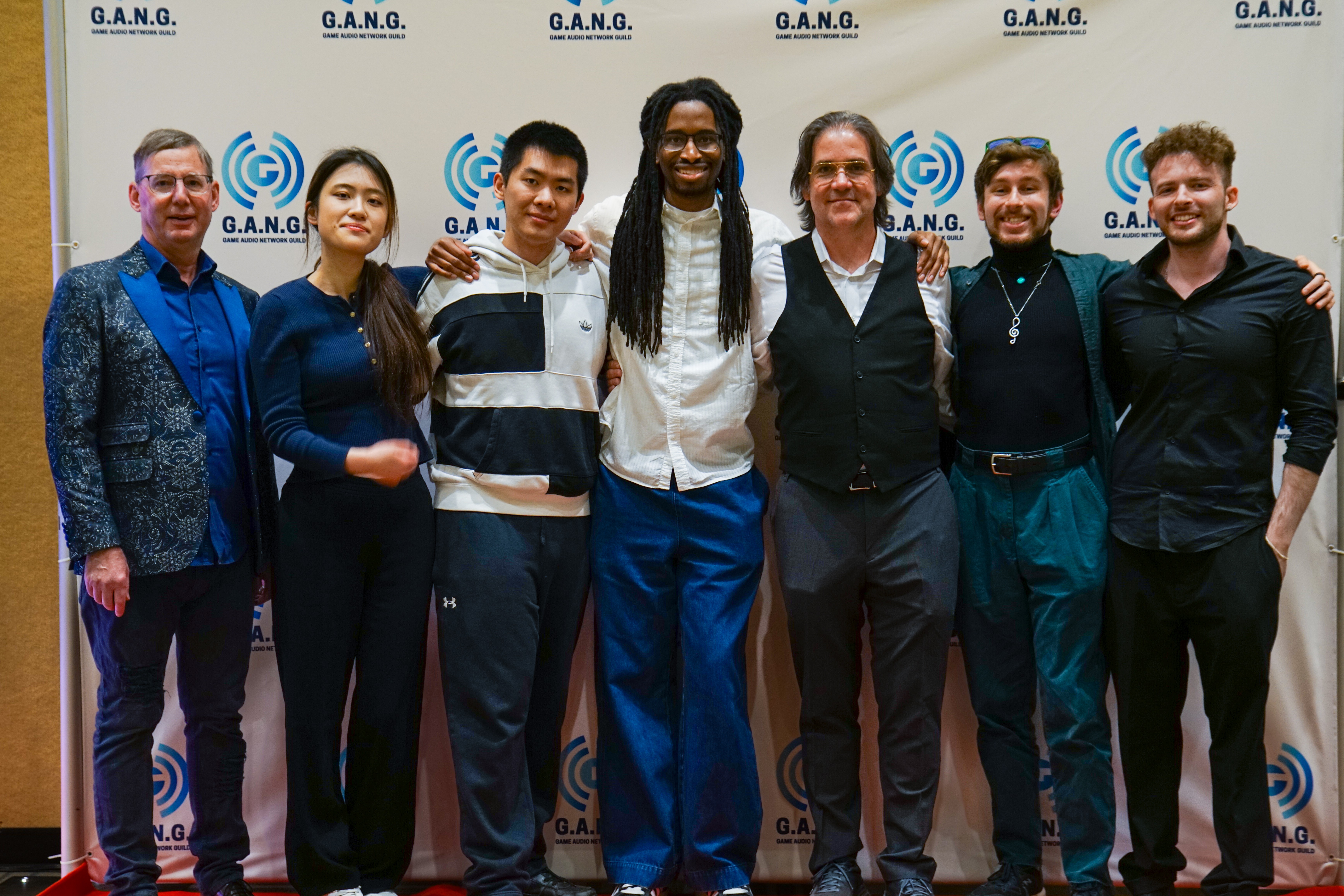 Group of seven diverse people standing together in front of a backdrop with logos, smiling, dressed in business casual attire.