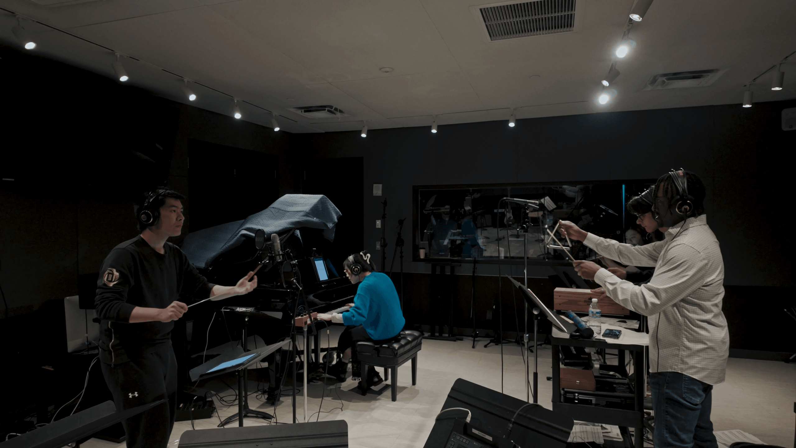 Three people in a recording studio with a person playing a keyboard, another reading sheet music, and a third person with a microphone.