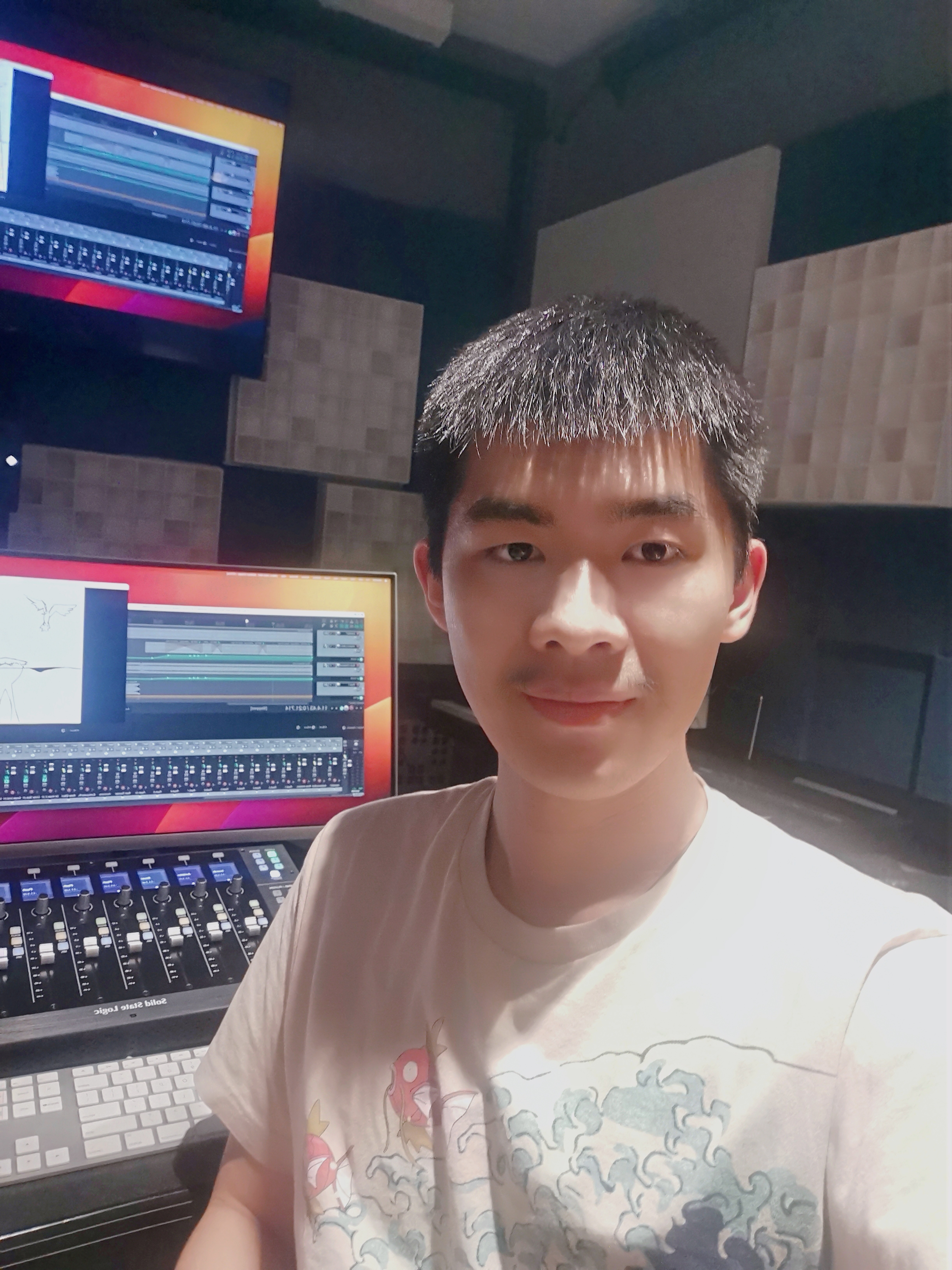 Young man with short black hair smiling in a recording studio with audio equipment and screens behind him.