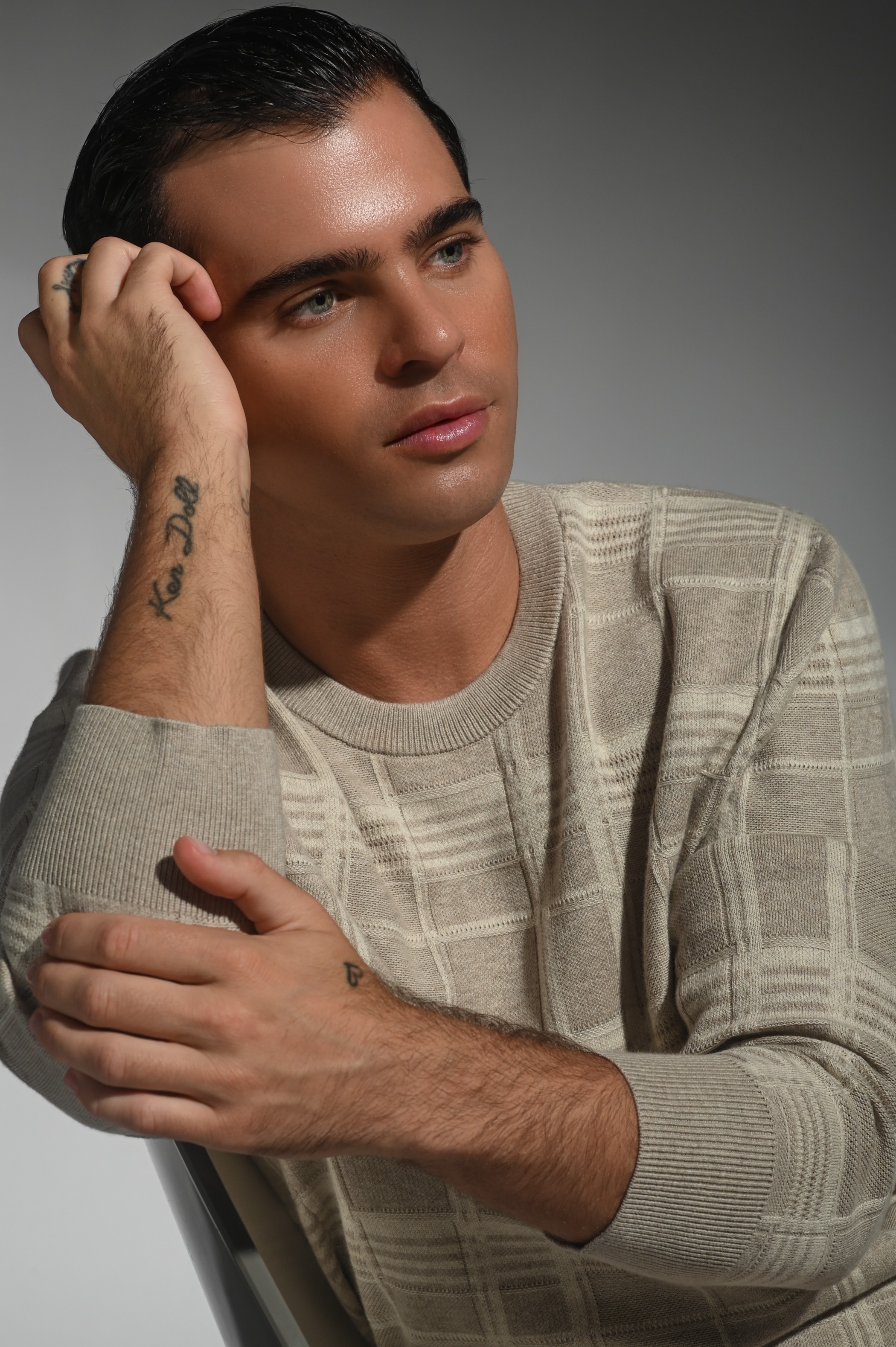 Young man with short dark hair, wearing a beige striped sweater, resting his head on his hand, looking thoughtful.