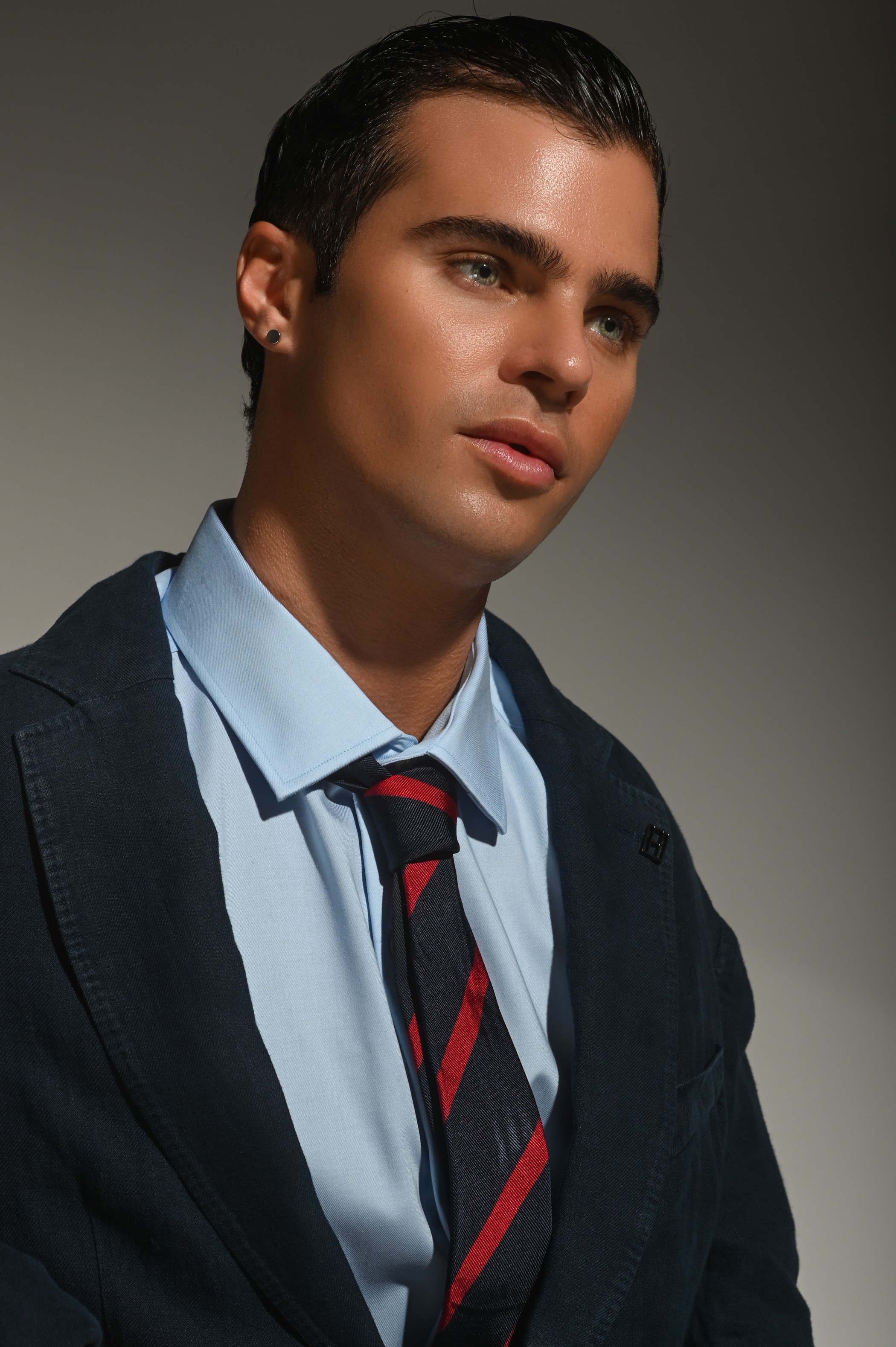 Young man with dark hair, wearing a light blue shirt, dark blazer, and a striped tie, looking to the side.
