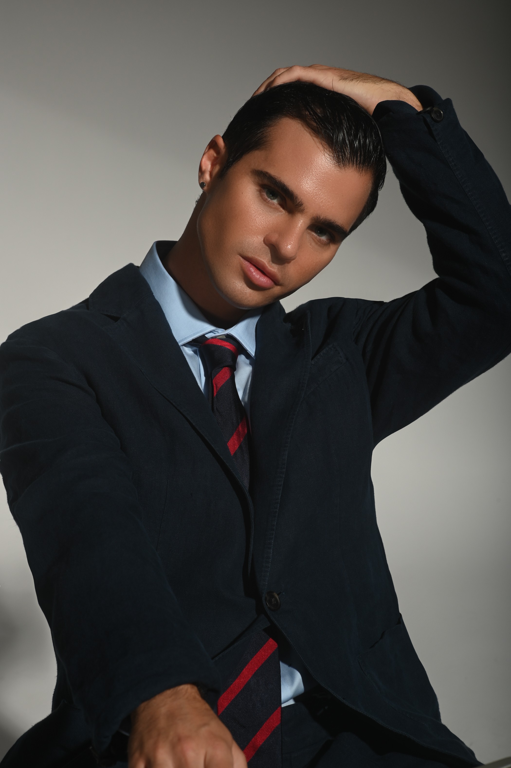 Young man with dark hair in a navy blazer, white shirt, and striped tie, posing with hand on head.