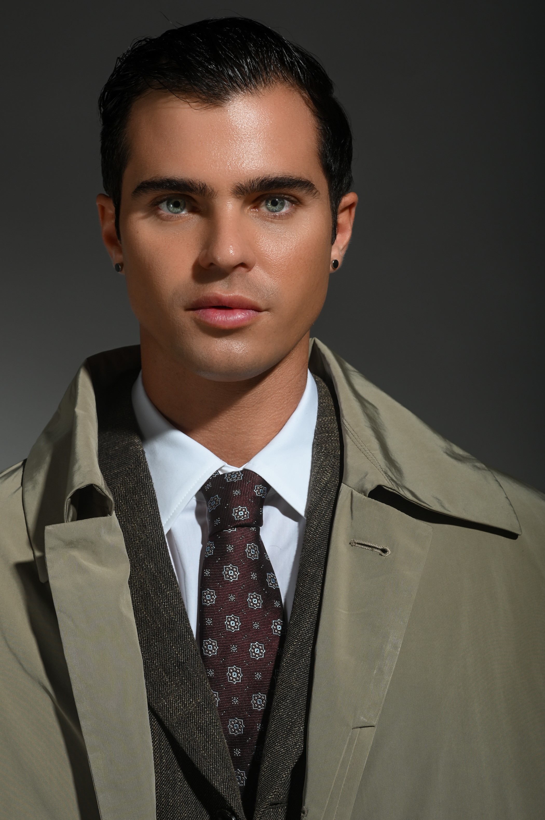 Portrait of a young man with dark hair, wearing a beige trench coat, white shirt, and patterned tie, against a dark background.