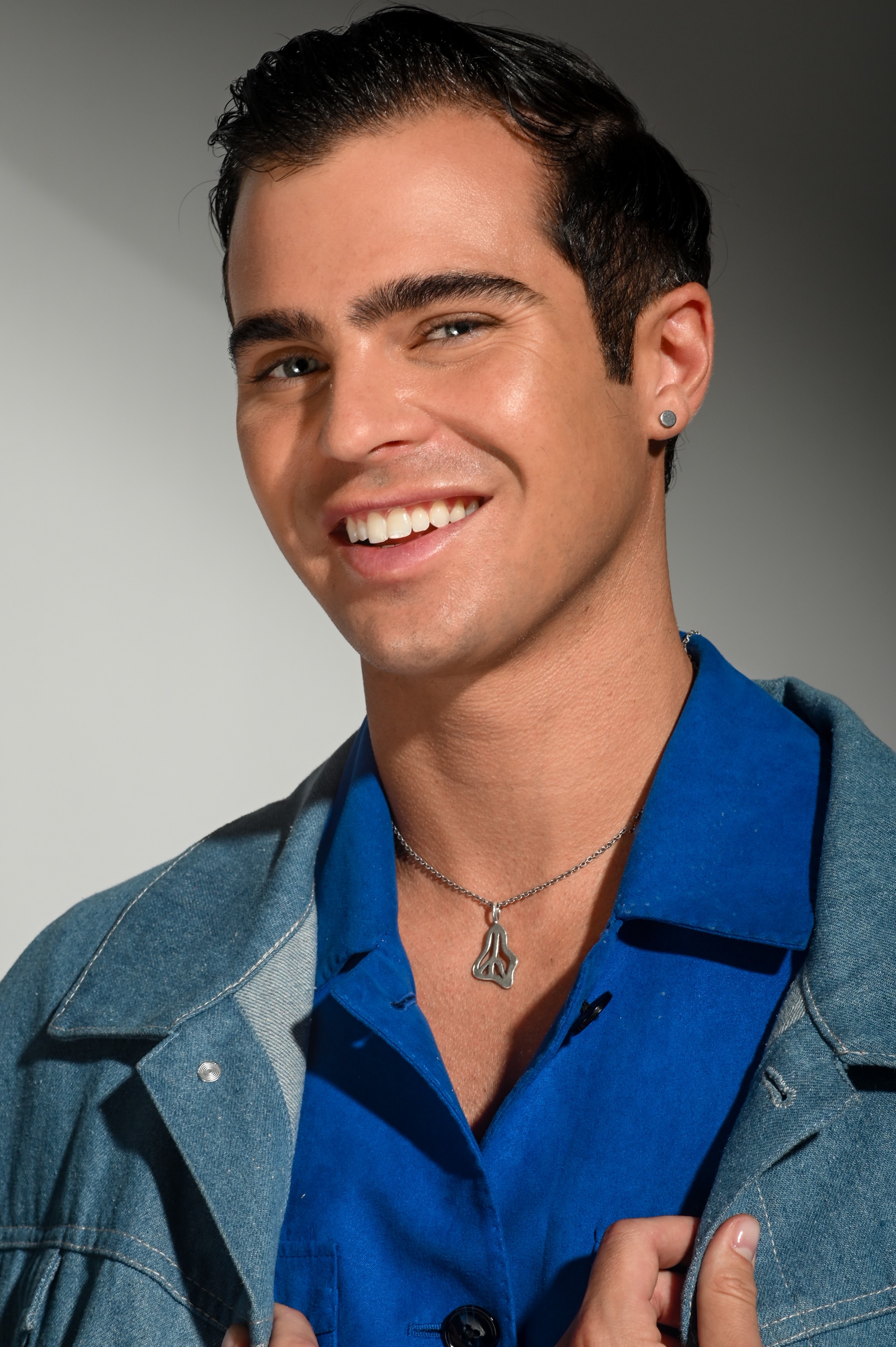 Young man with dark hair, smiling, wearing a blue shirt and denim jacket, against a neutral background.