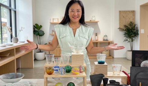 Woman smiling with arms outstretched in a bright, modern room with shelves, plants, and tableware displays.