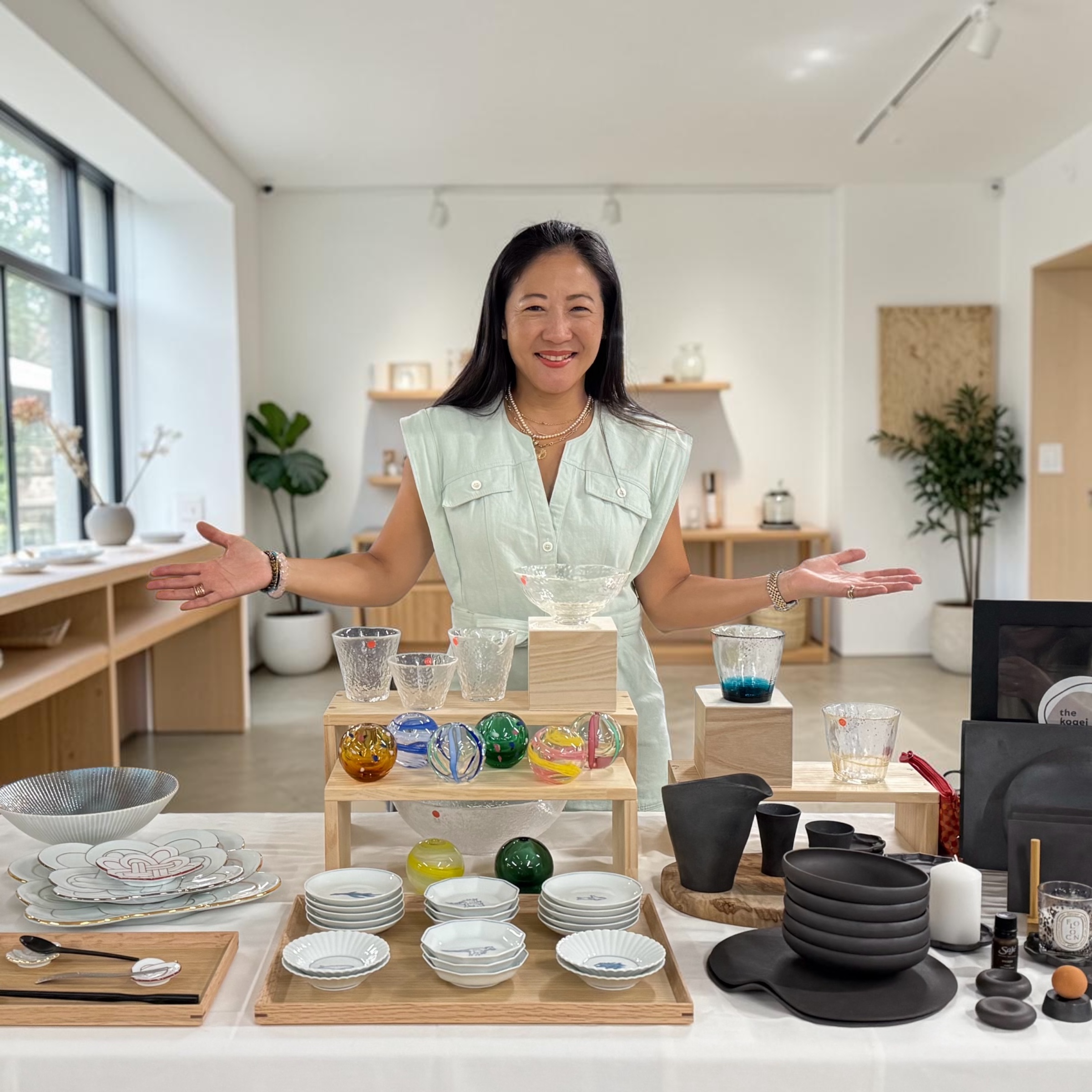 Woman smiling with arms outstretched in a bright, modern room with shelves, plants, and tableware displays.