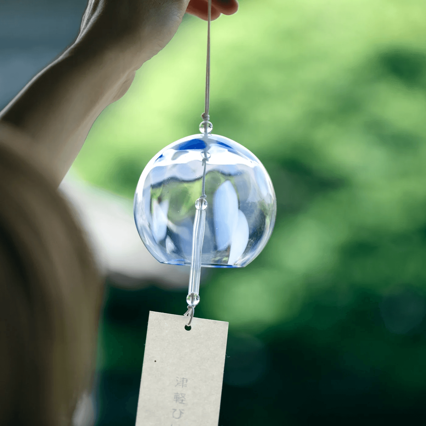 Person holding a glass wind chime with a tag, outdoors with green blurred background.
