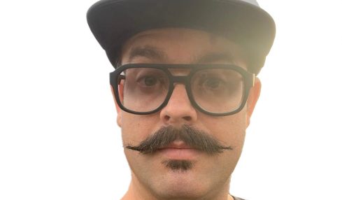Person with glasses, mustache, wearing a cap, and a dark shirt, looking at the camera against a white background.