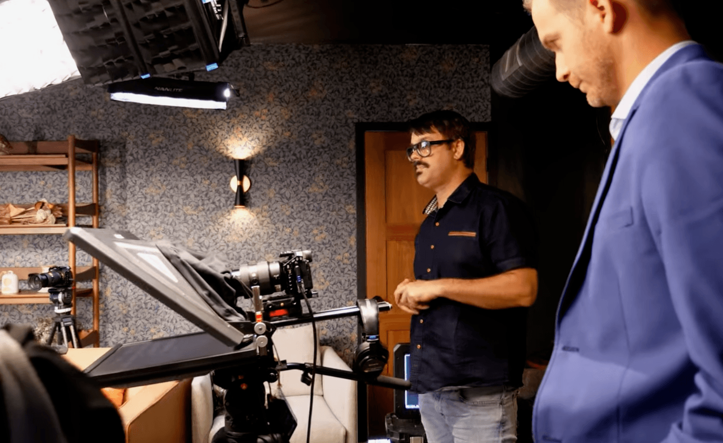 Two men in a room with professional camera equipment, one in a suit and the other in casual clothing, preparing for filming.