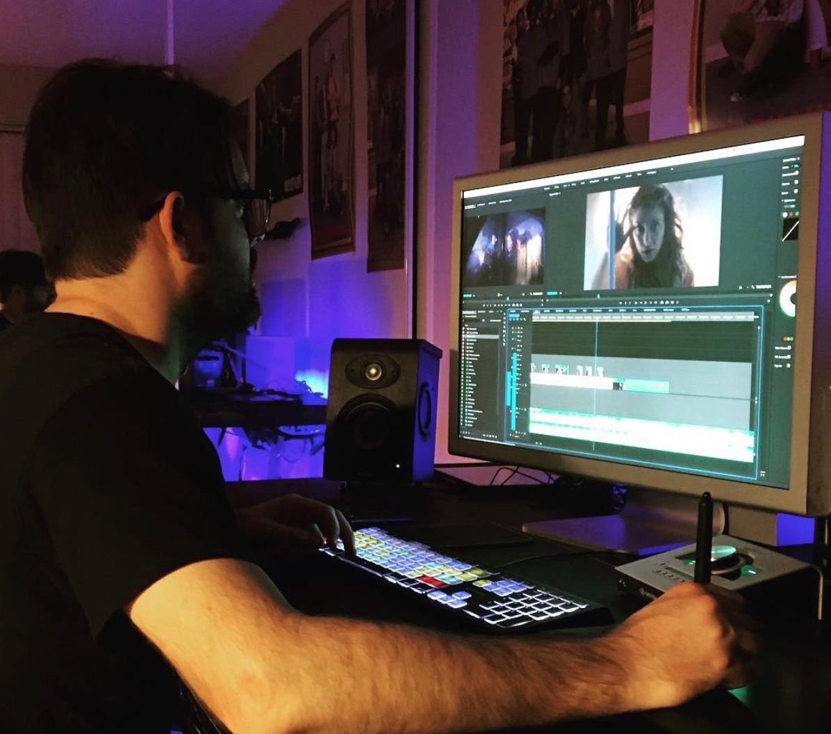 Person working on video editing at a computer with a large monitor, in a dimly lit room with framed pictures on the wall.