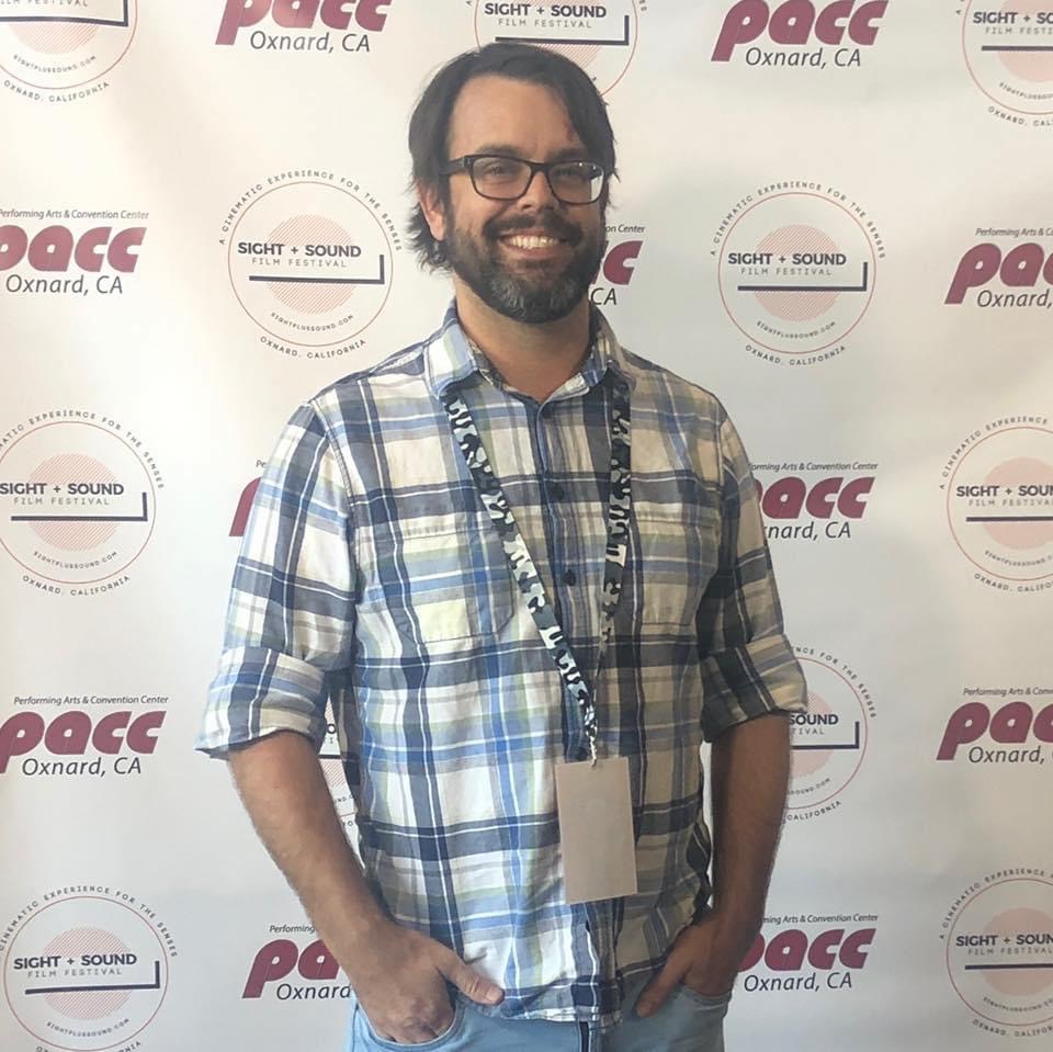 Man with glasses and beard wearing a plaid shirt, smiling, standing in front of a backdrop with logos.