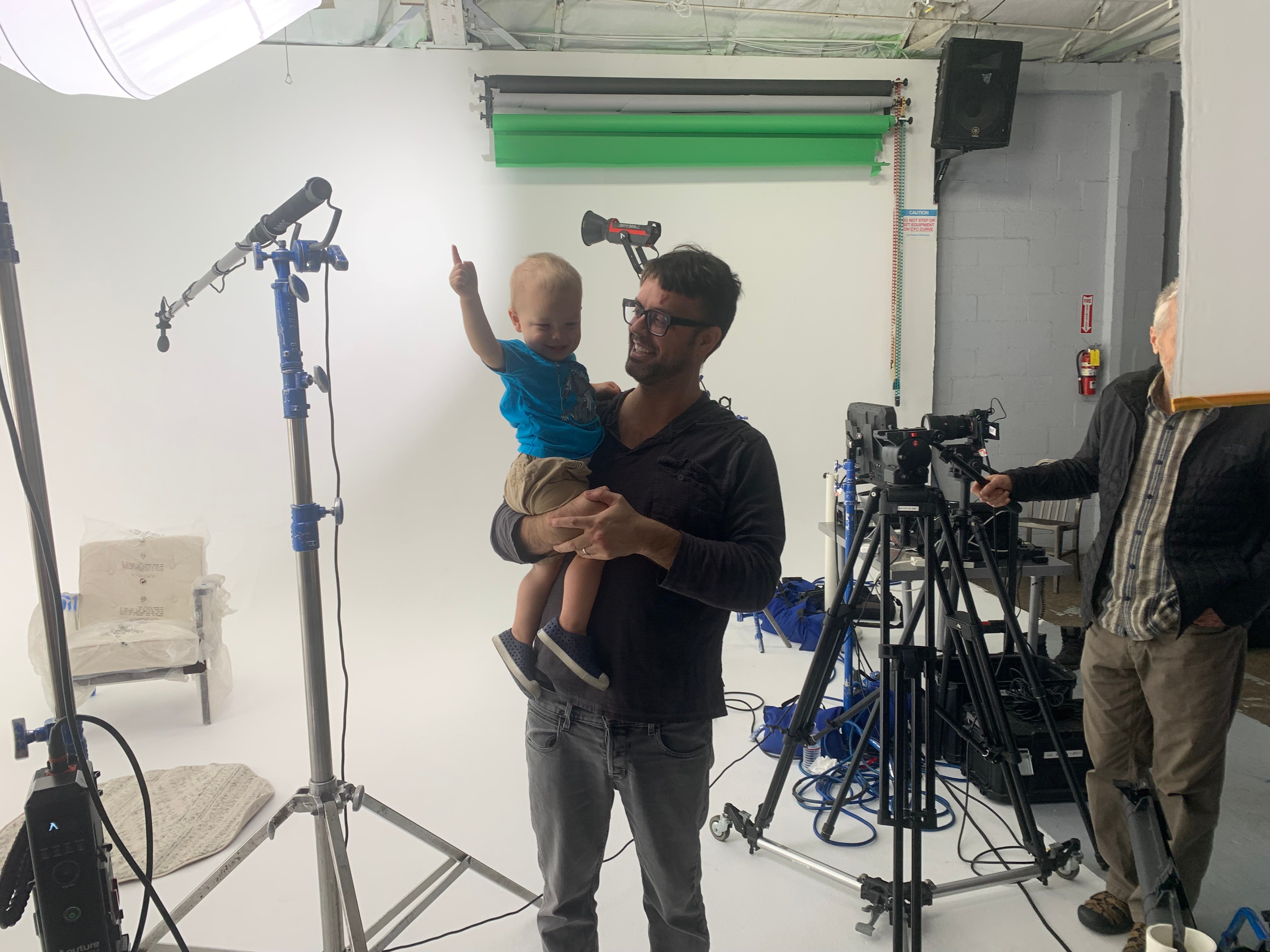Man holding a child in a studio with cameras and lighting equipment, green backdrop, and a white chair.