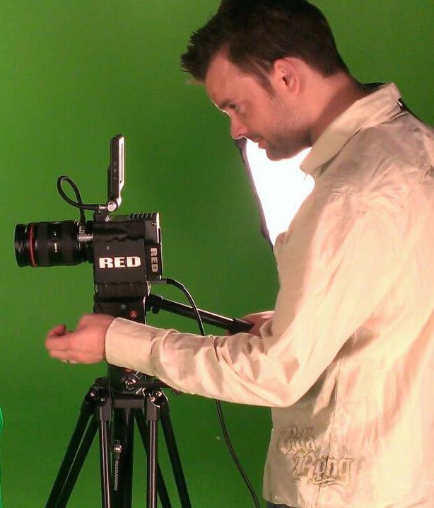 Man operating a camera on a tripod against a green screen, looking down at the equipment.
